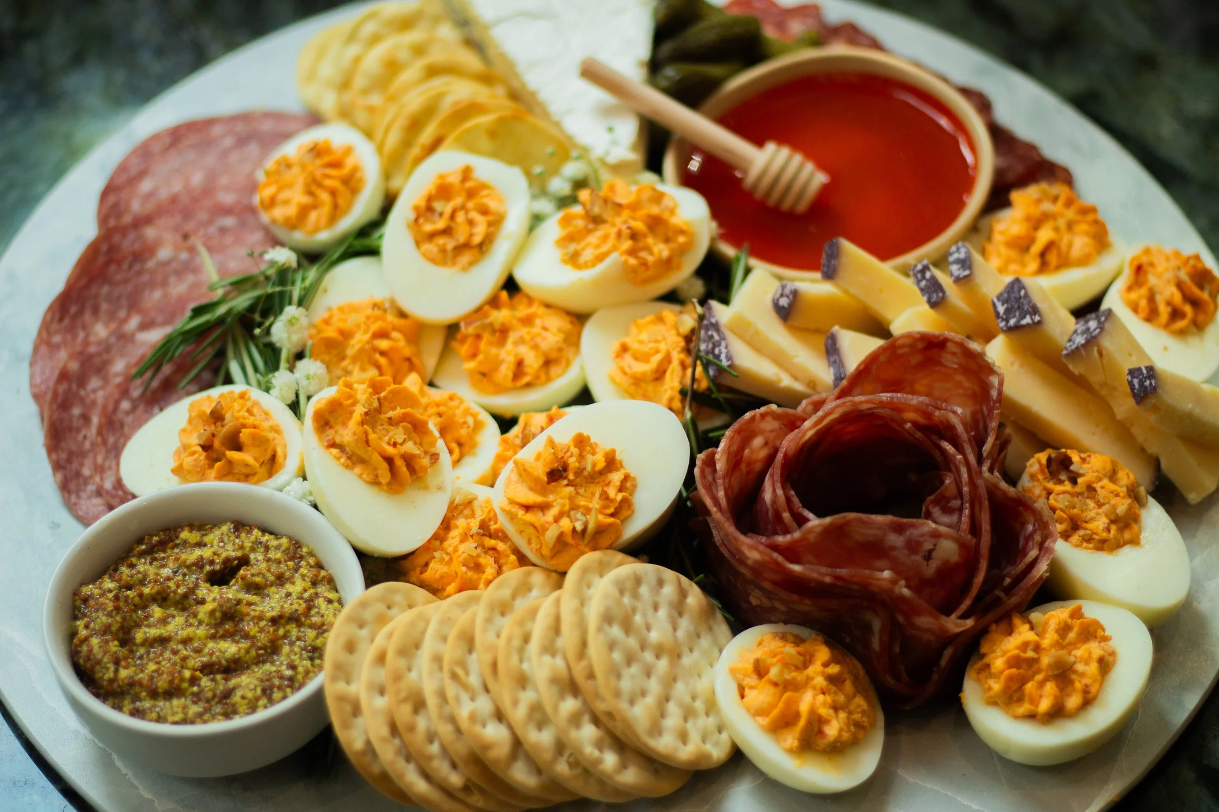A cheese and meat platter with sliced hard-boiled eggs topped with spicy filling, slices of salami, and cheese sticks, served with mustard, honey, and tomato sauce.