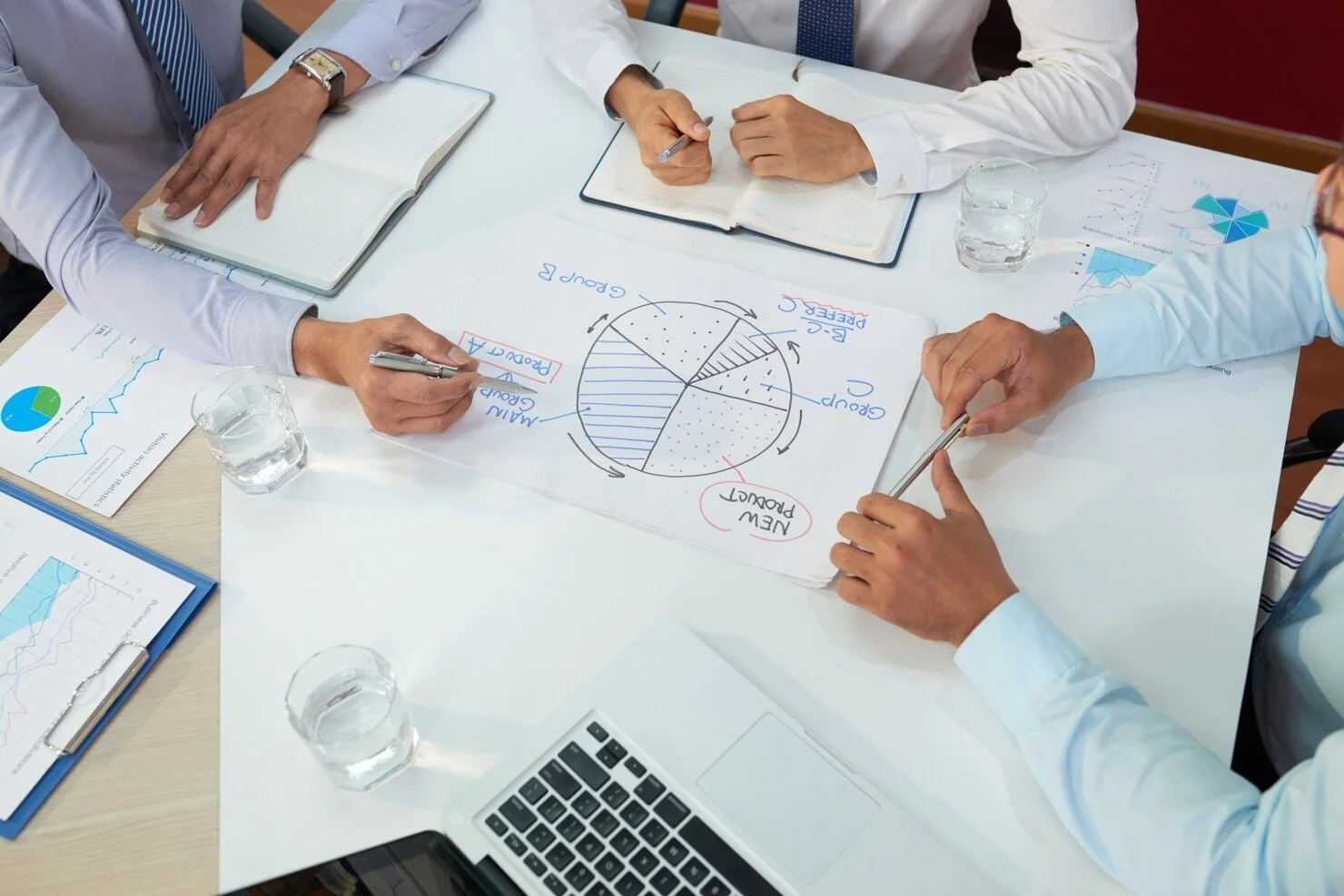 Business meeting with four professionals reviewing a pie chart and diagrams on paper, surrounded by notebooks, glasses of water, and charts, in a conference room.