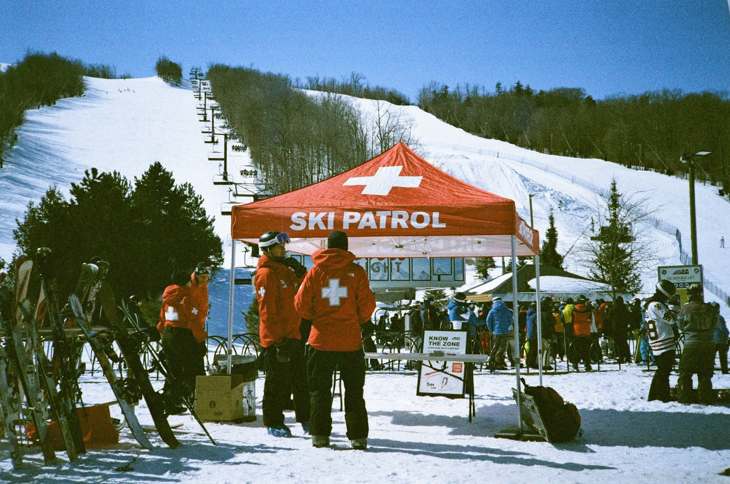 Ski patrol tent on a snowy mountain with people in winter gear, ski racks, and ski lifts in background, under clear blue sky.