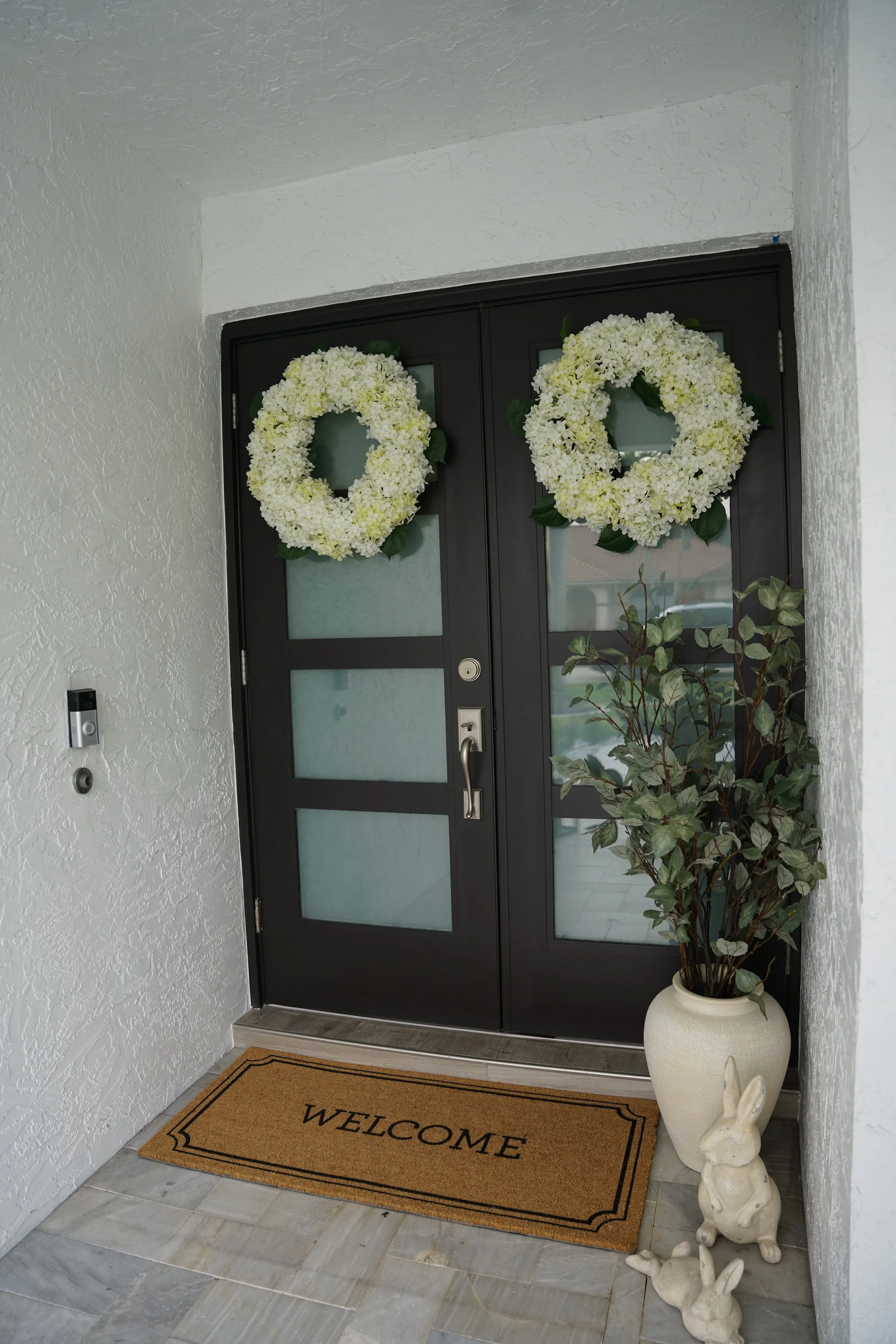 Black front door decorated with two white floral wreaths, with a welcome mat in front, and potted plants and bunny statues on the side.