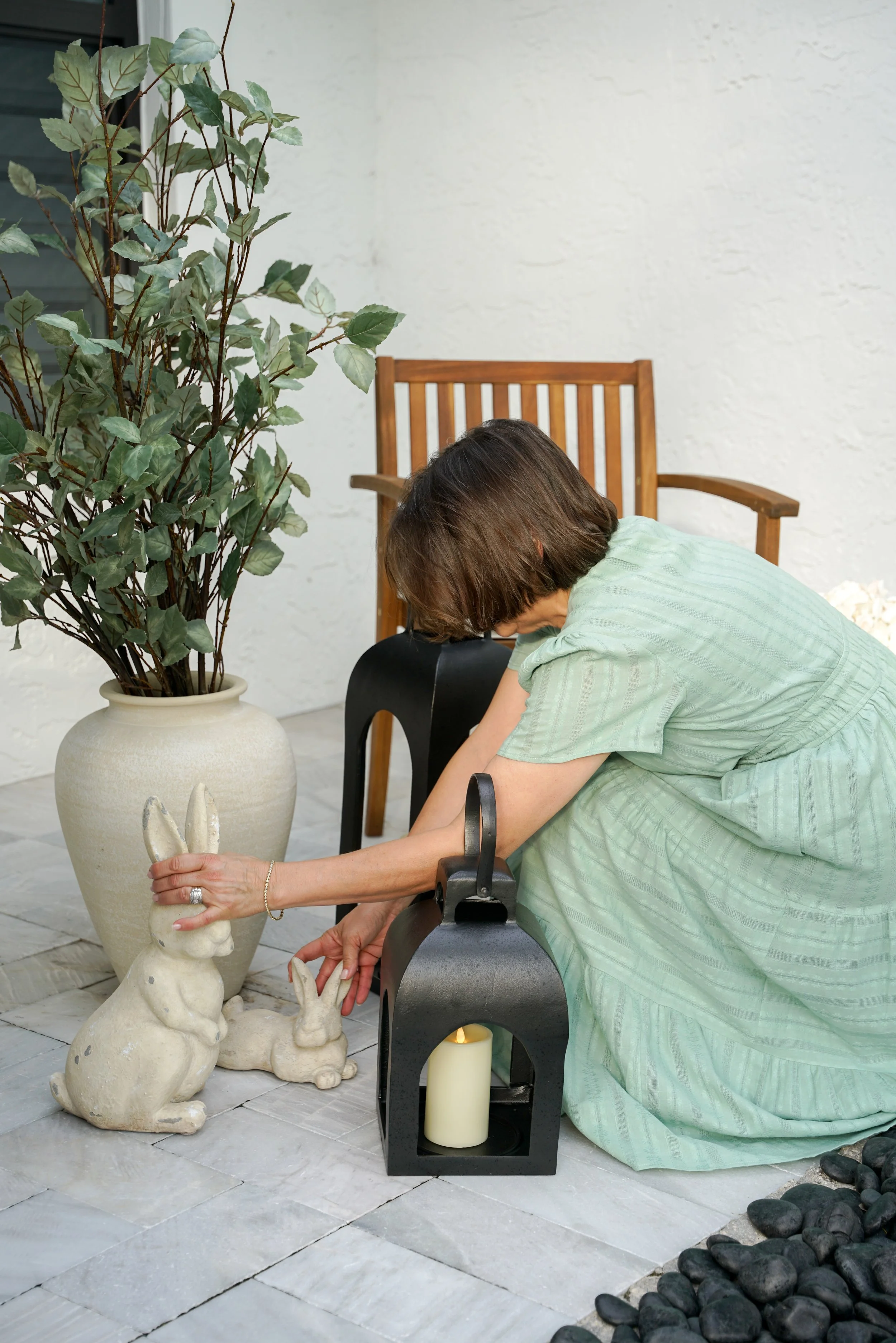 A woman in a green dress arranging two ceramic bunny garden statues on the porch. There is a large potted plant with green leaves and a white wall in the background, along with a black lantern and a battery-operated candle next to her.