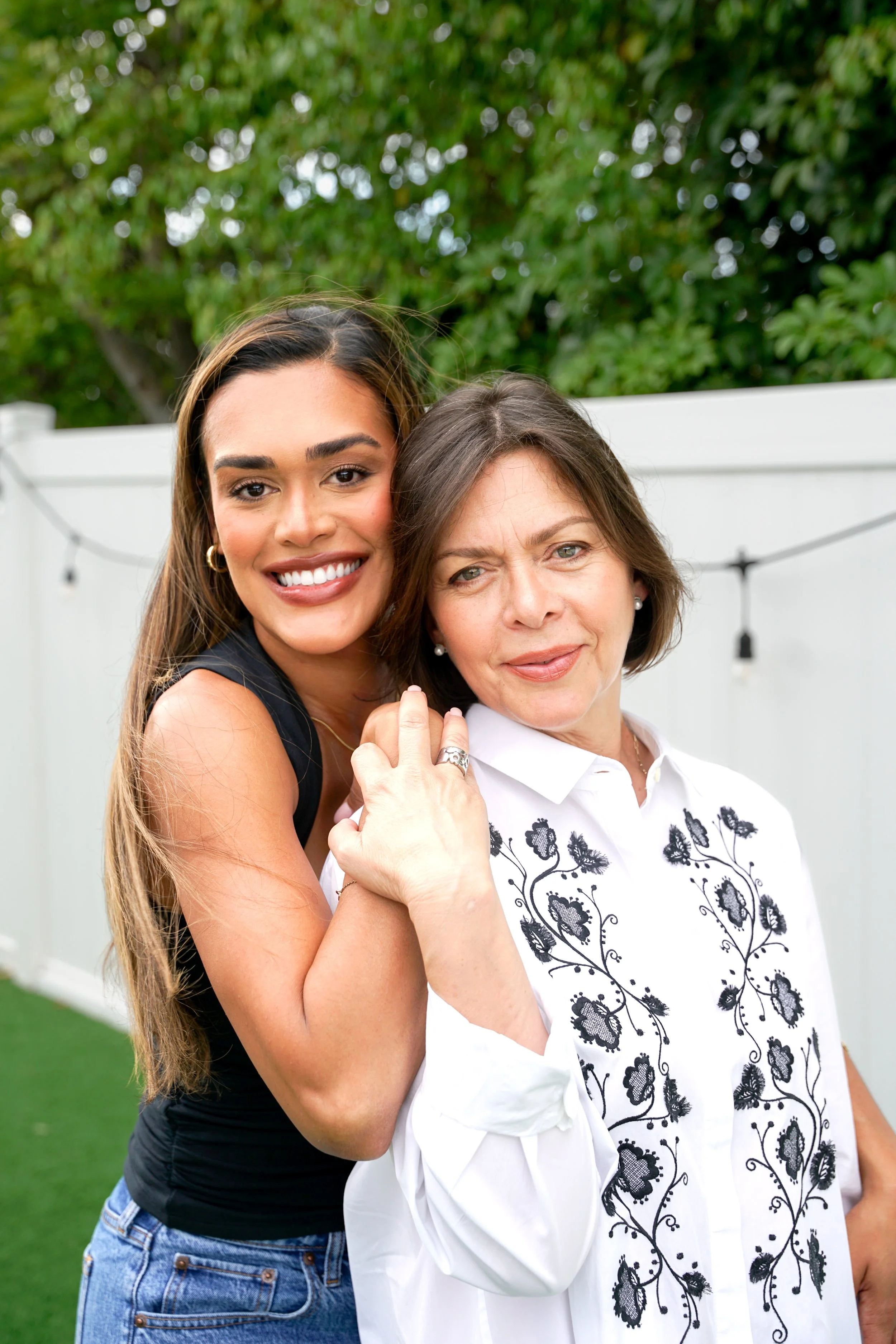 Two women embracing outdoors, smiling at the camera, standing in front of a white fence and green trees.