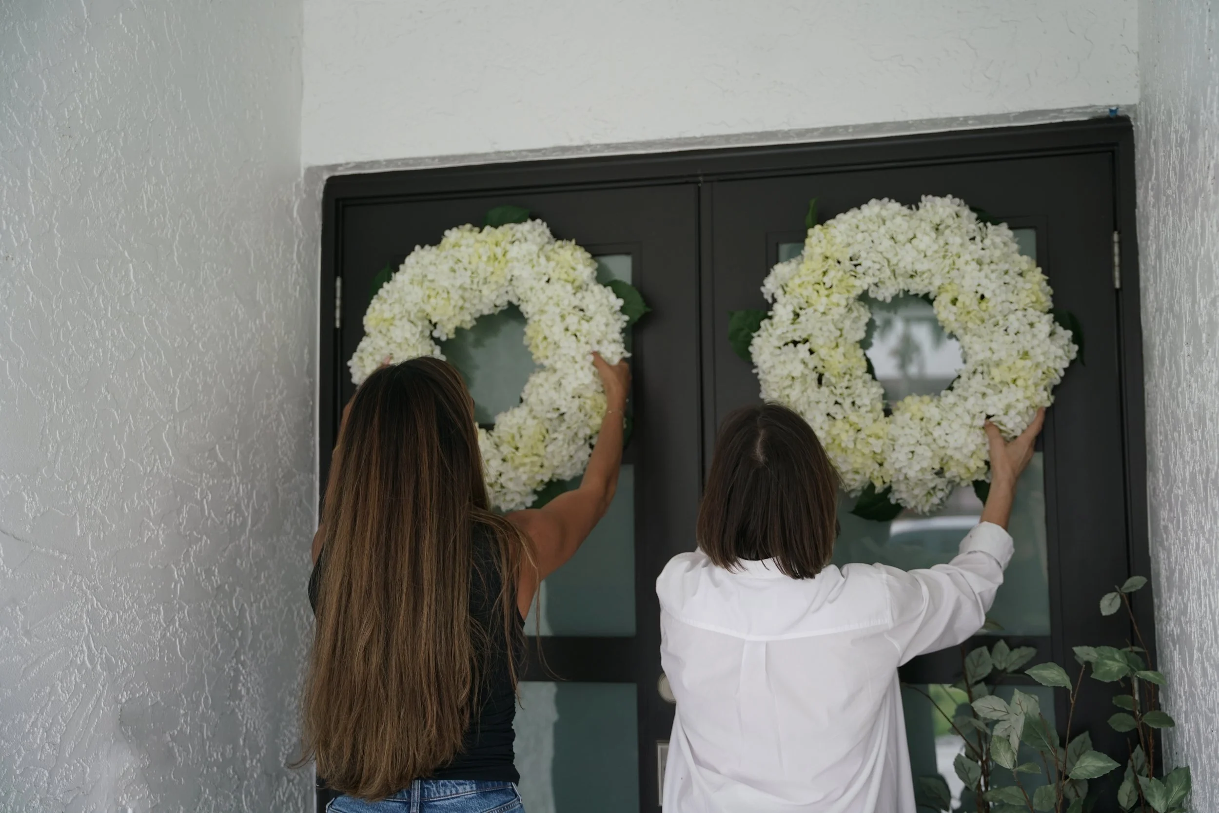 Two women hang white floral wreaths on a black door decorated for a celebration.