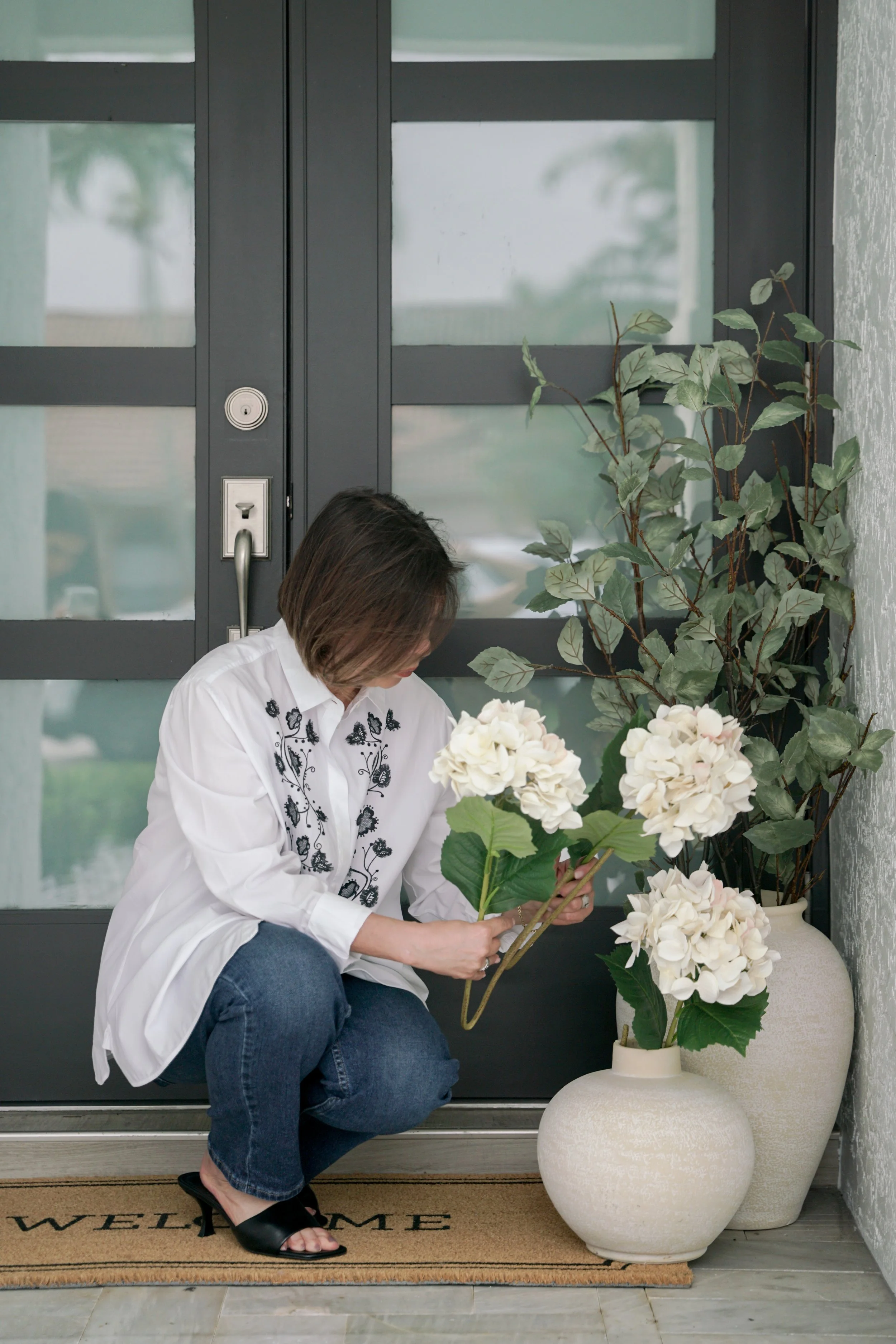 A woman with short dark hair, wearing a white embroidered blouse, crouches by the door and arranges a bouquet of white hydrangea flowers in a white ceramic vase, with a large plant beside her on the right.