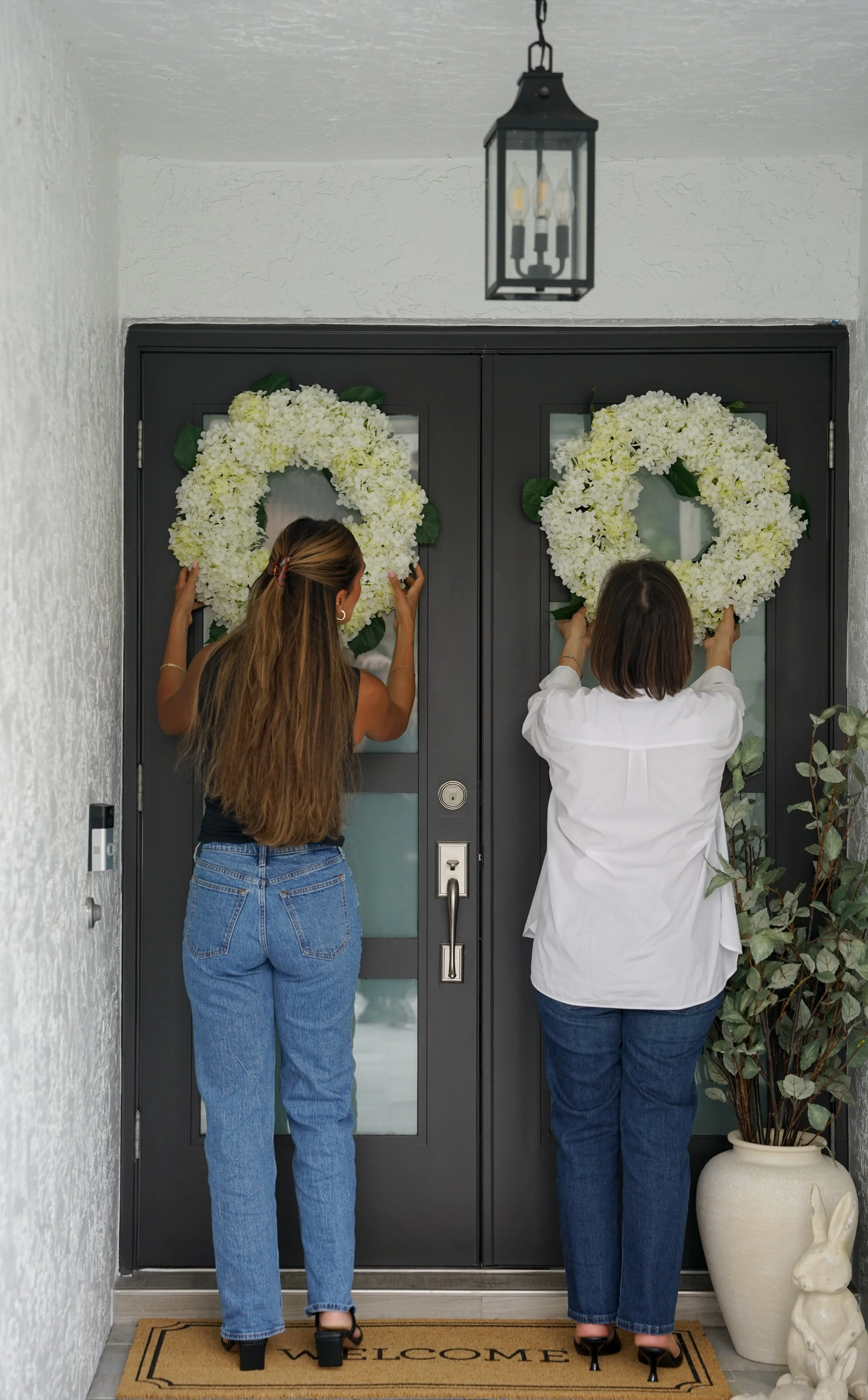 Two women are placing white floral wreaths on a door at the entrance of a home. The woman on the left has long hair and is wearing a black top and blue jeans. The woman on the right has shorter hair and is dressed in a white blouse and dark jeans. There is a welcome mat on the floor and a large ceramic vase with greenery on the side.