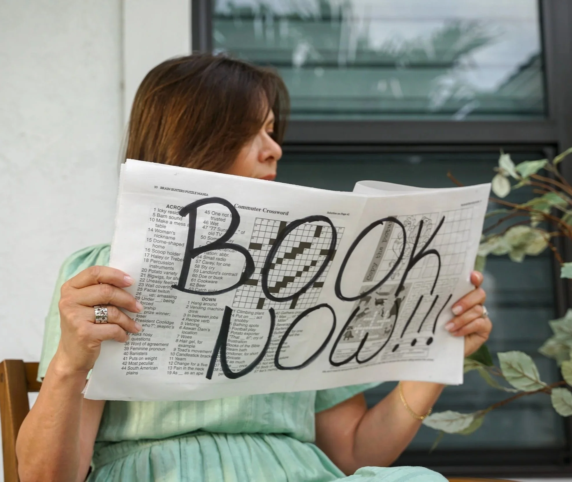 Woman reading newspaper with large handwritten message 'BOO!'.