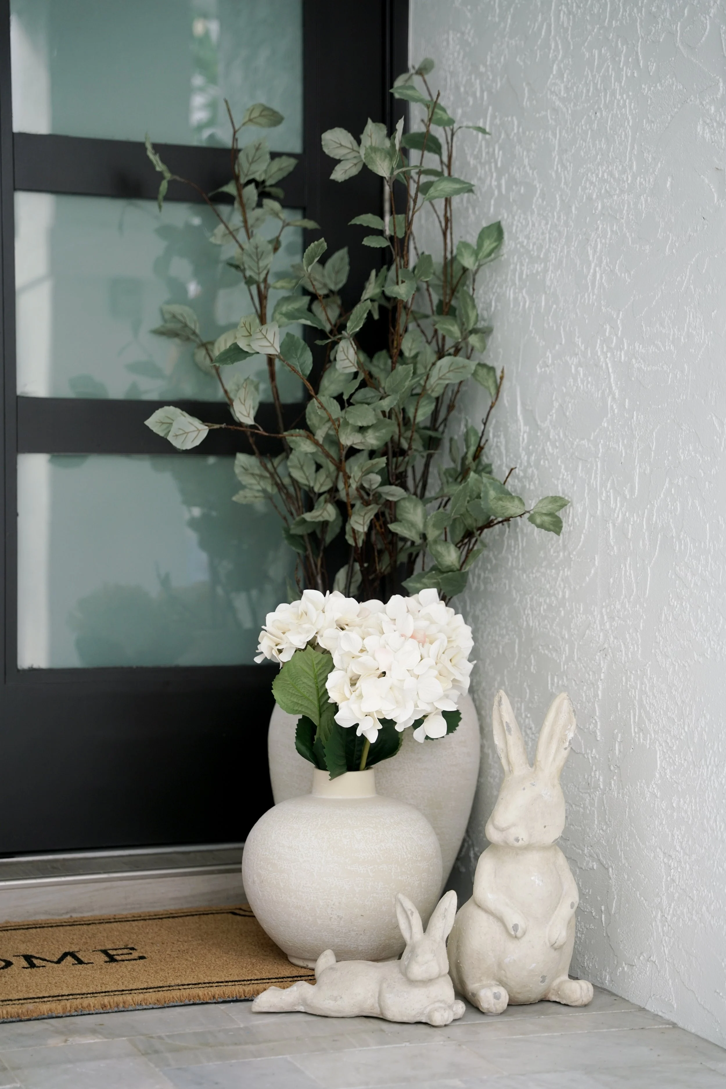 Decorative white vases with artificial white hydrangea flowers and bunny statues near an entry door with a doormat that says 'Welcome'.