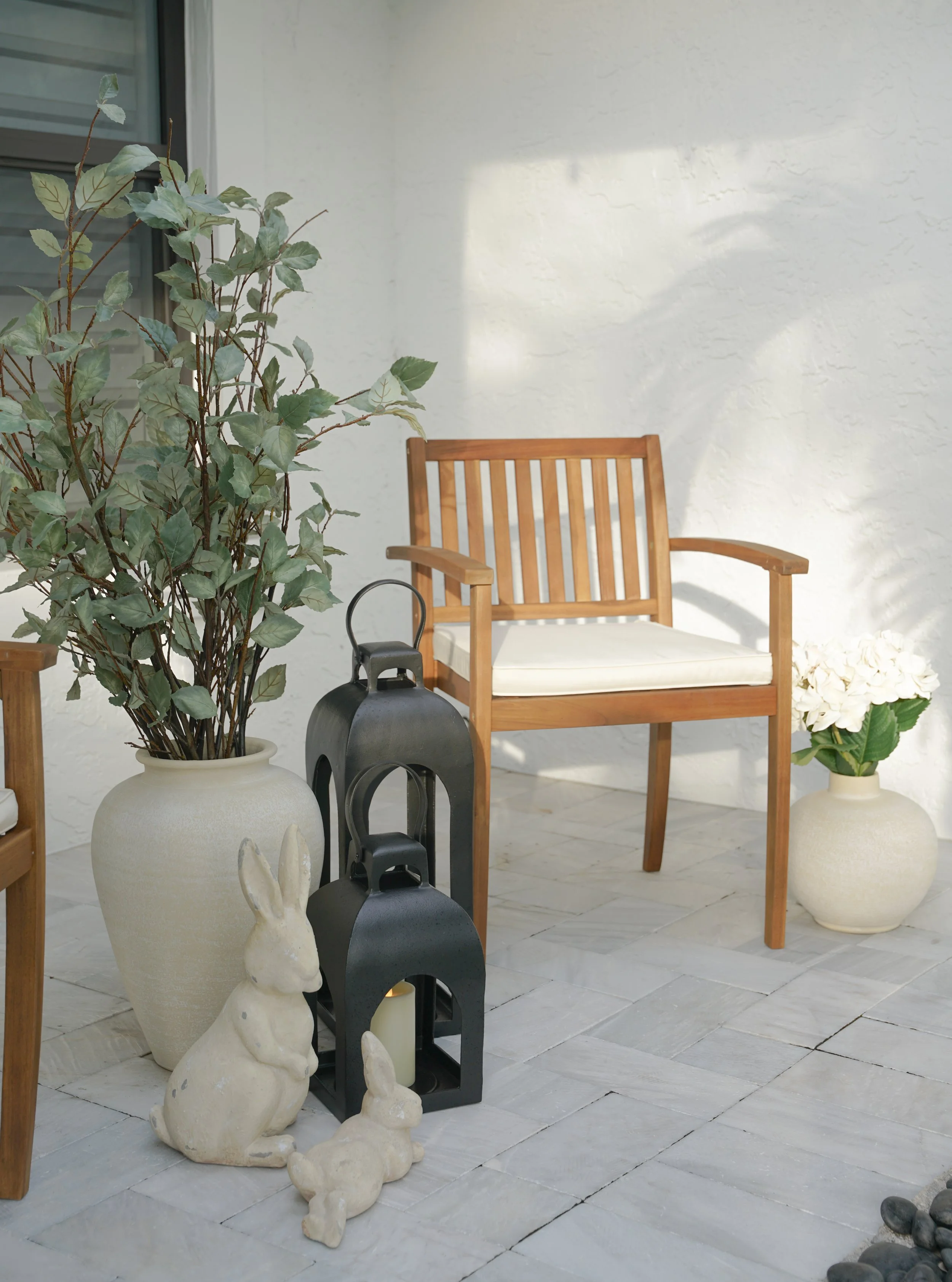 Decorative patio setup with potted plants, a wooden bench with cushion, bunny-shaped ornaments, and lanterns with candles on a stone-tiled floor.