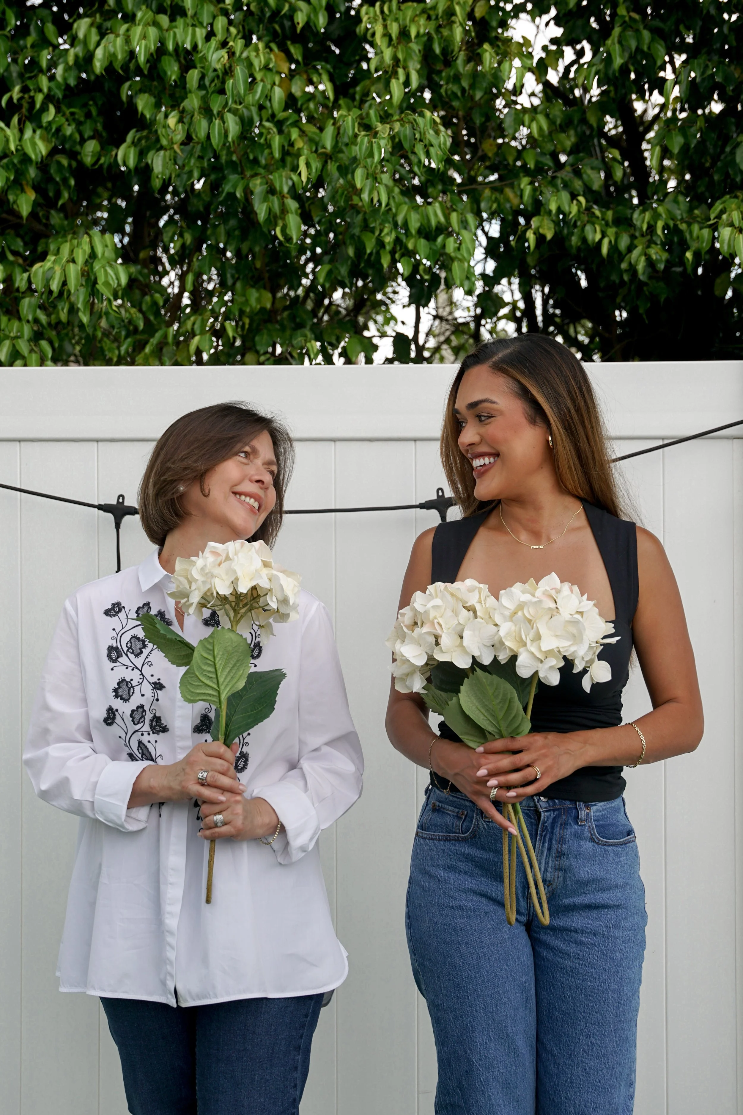 Two women smiling and holding bouquets of white hydrangeas in a backyard with a white fence and green tree.