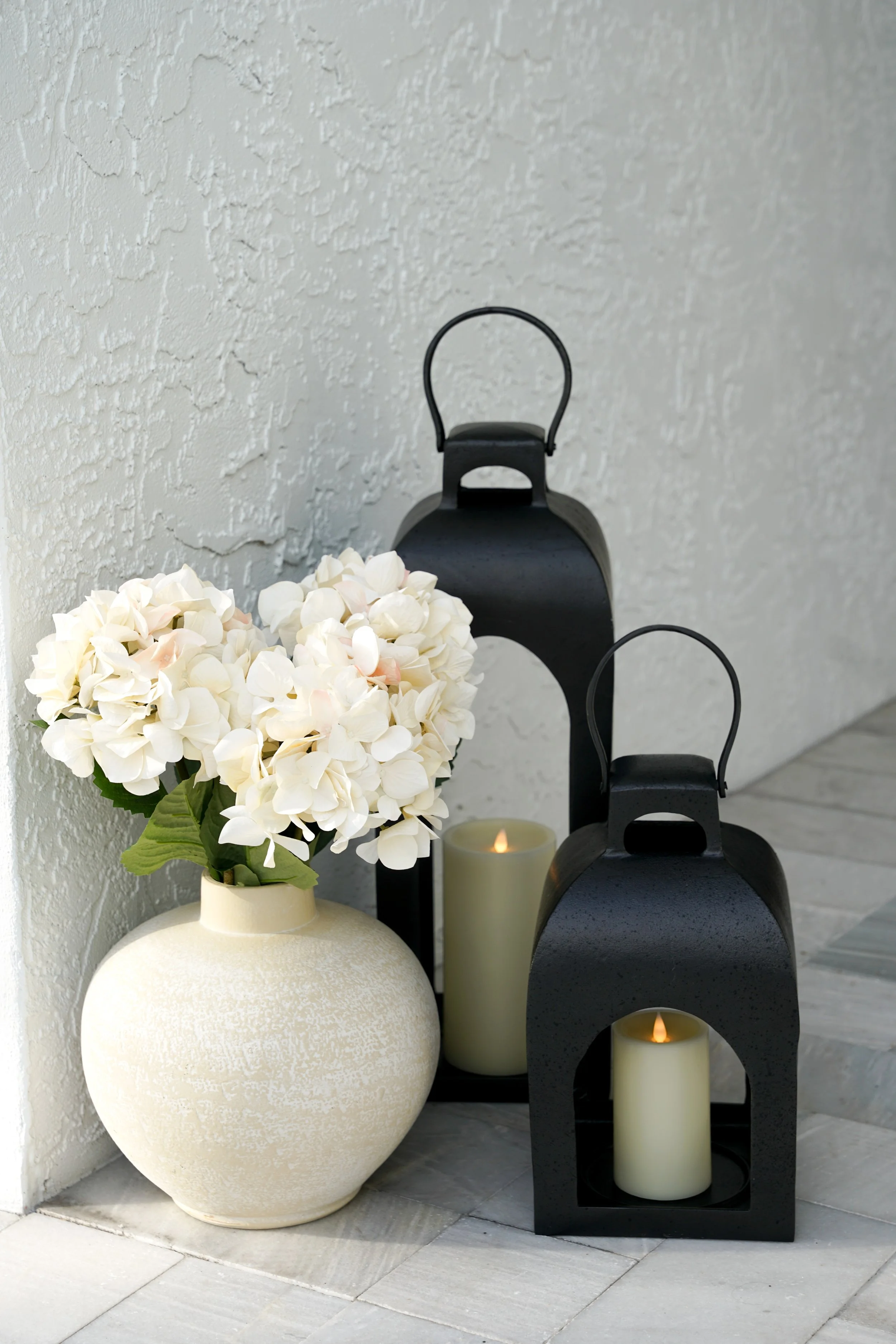 A decorative arrangement with a cream-colored vase holding white hydrangea flowers, flanked by two black lanterns containing candles, placed on a tiled floor against a textured white wall.