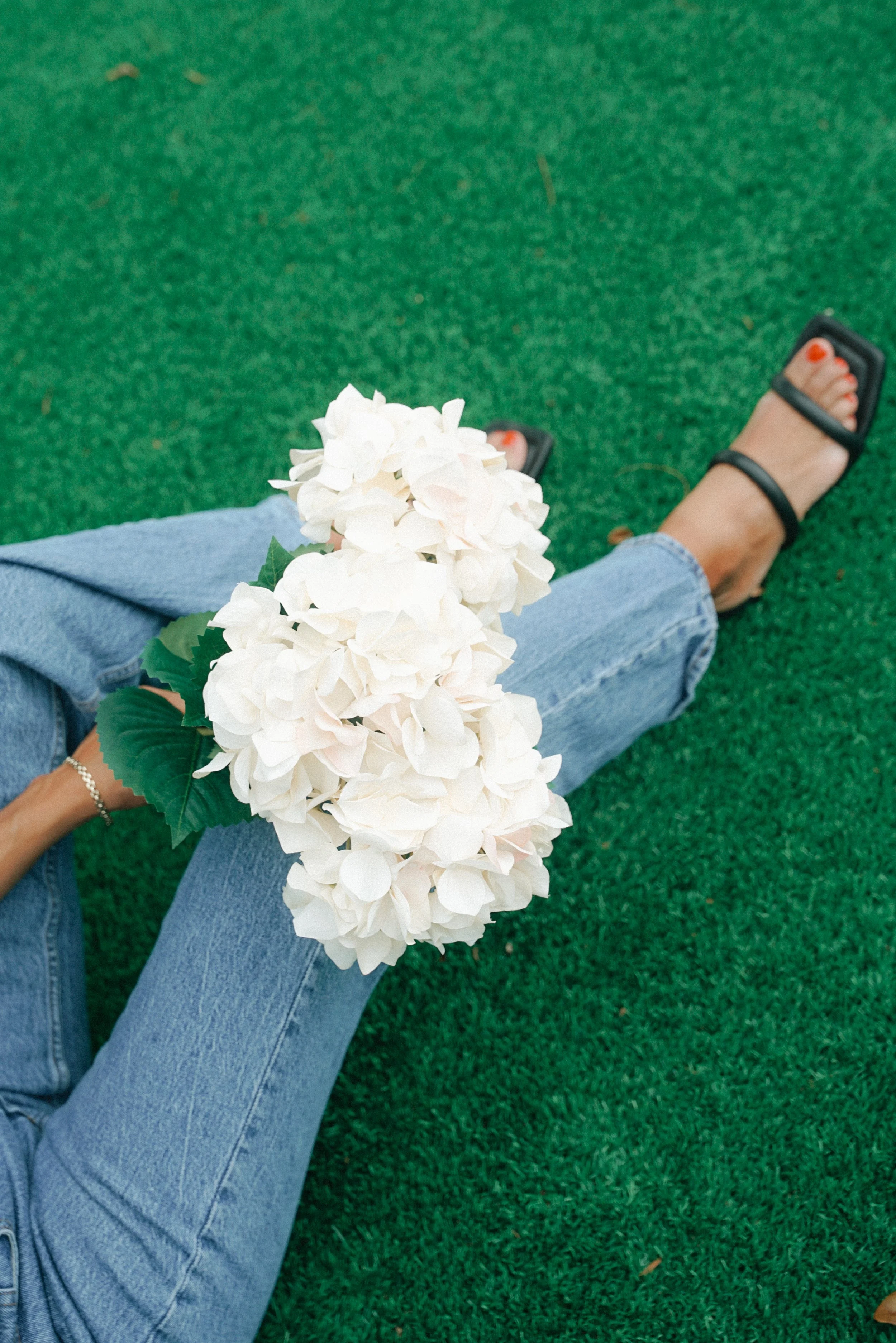 Person sitting on green grass holding a bunch of white hydrangea flowers, wearing light blue jeans and black sandals with red painted toenails.