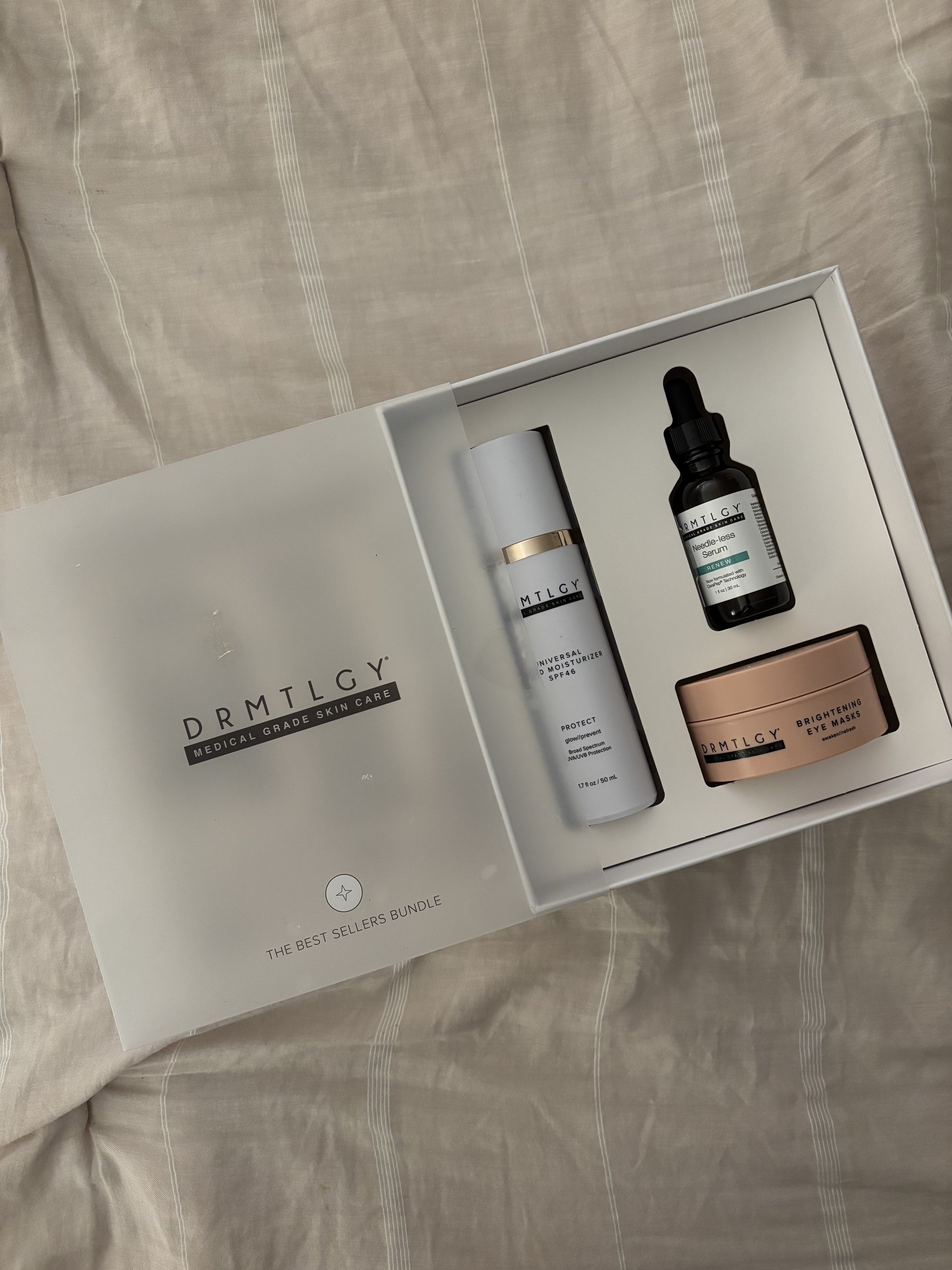 Drugstore skincare set with a moisturizer, serum, and eye masks from DRMTLGY in a white box on a beige bed.
