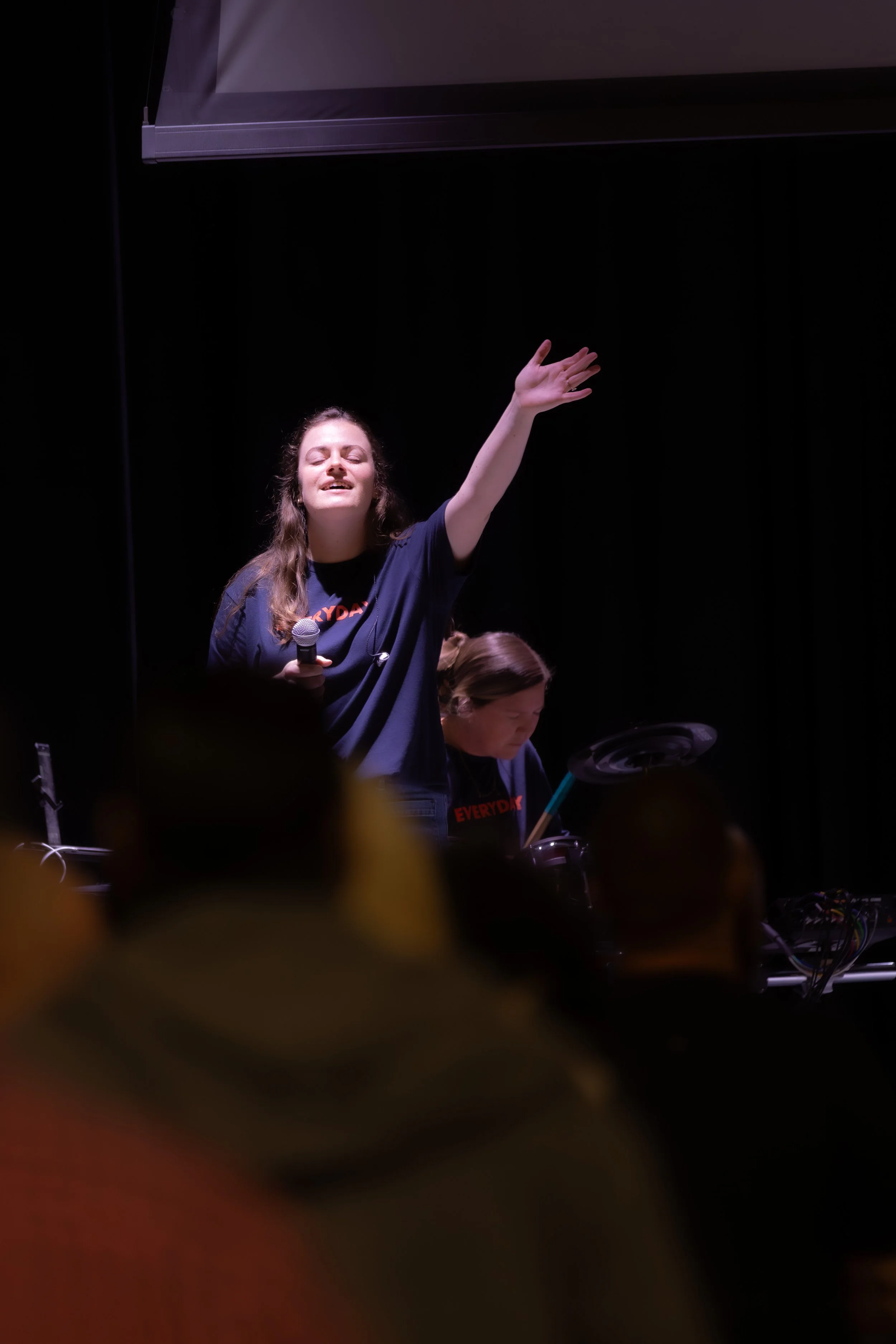 A woman on stage with long wavy hair, eyes closed, smiling, holding a microphone in her right hand, and raising her left hand. She is wearing a dark T-shirt with orange text. Another woman in the background is focused on an electronic device. The stage has dark curtains and equipment visible in the foreground.