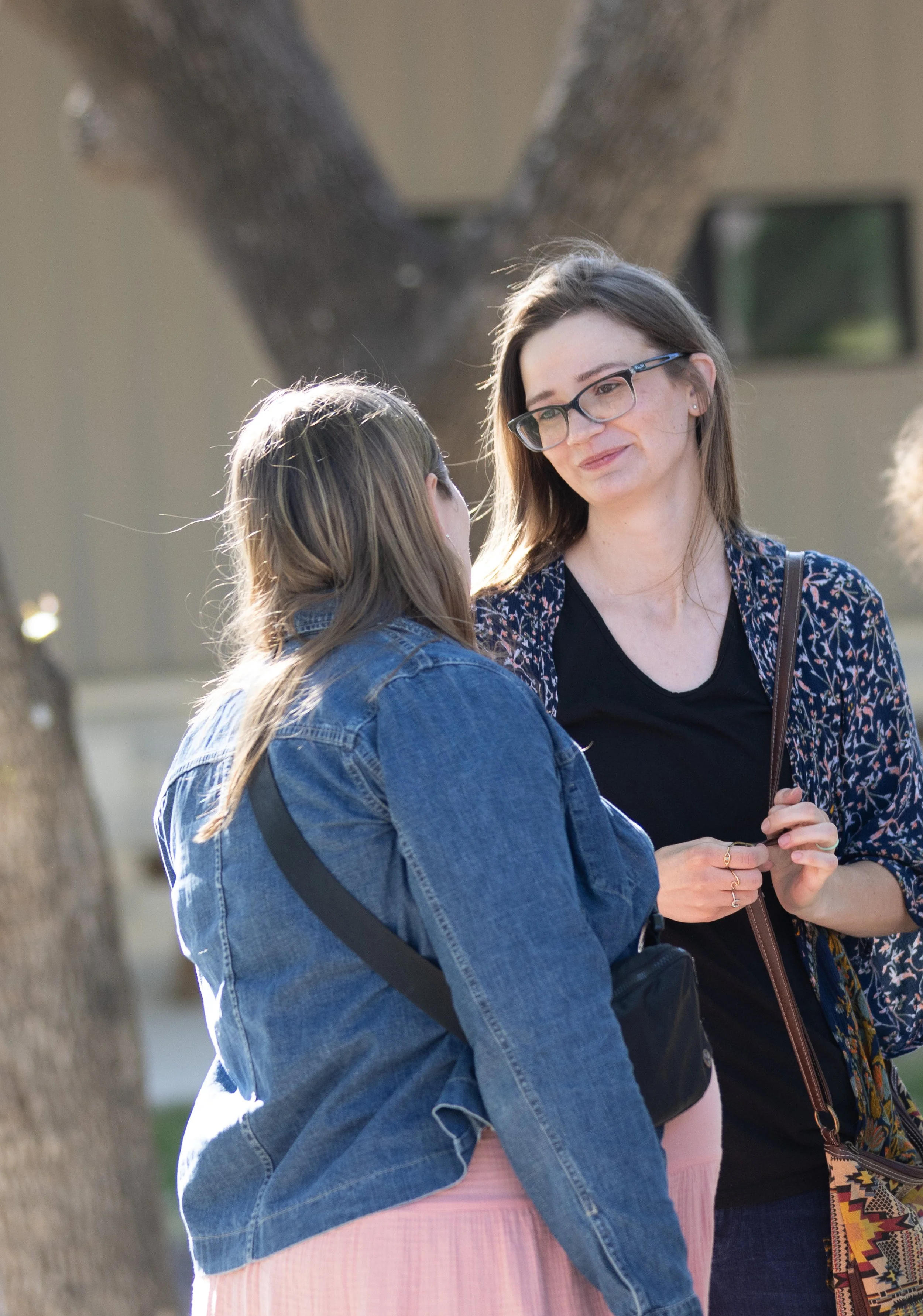 Two women having a conversation outdoors, one wearing glasses and a patterned jacket, the other with long brown hair in a denim jacket and pastel pink dress.