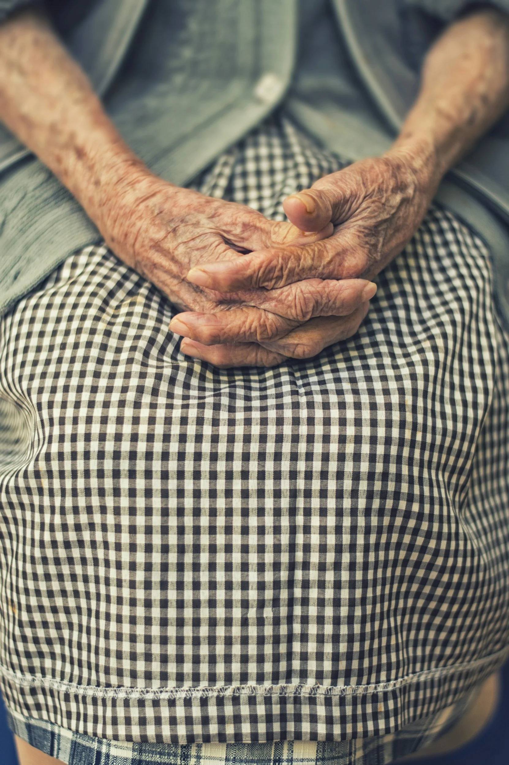 Close-up of elderly person's wrinkled hands resting folded on lap, wearing checkered pants and a denim jacket.
