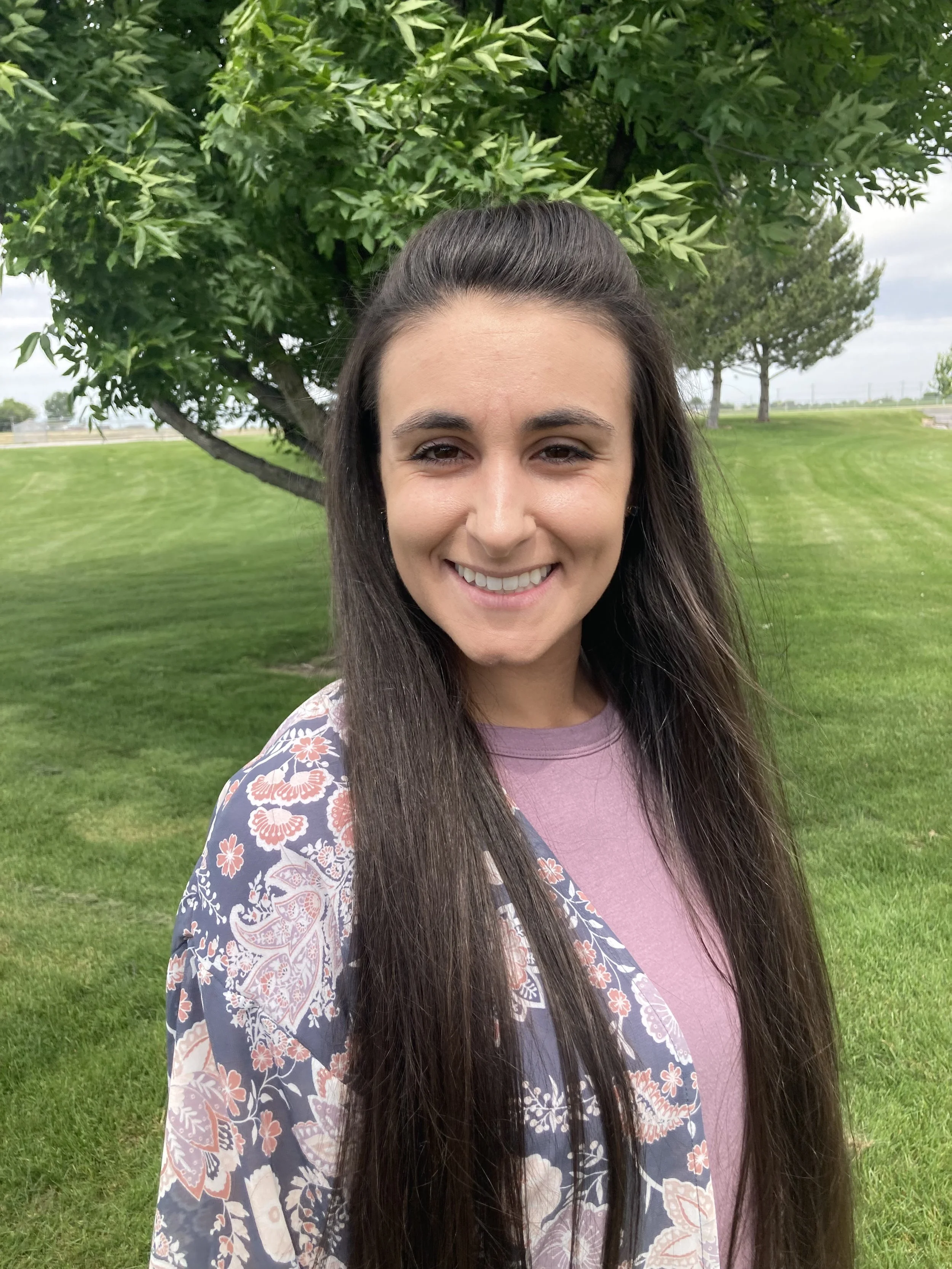 A woman with long dark hair smiling outdoors in front of a tree and green grass.