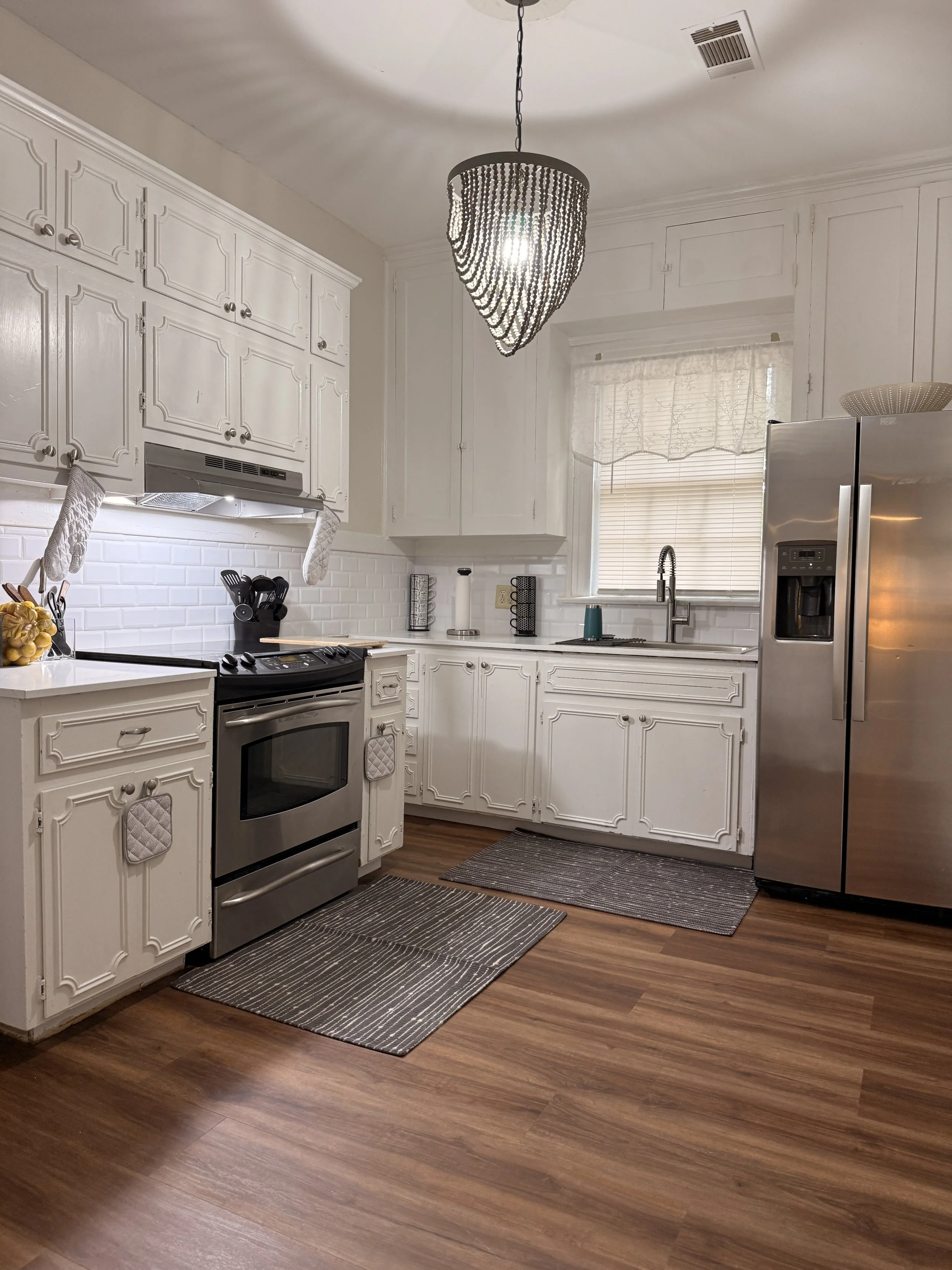 A white kitchen with white cabinets, stainless steel oven, and refrigerator. A black chandelier hangs from the ceiling, and there are two small rugs on the wooden floor. The kitchen has a window above the sink with a white curtain, and the countertop