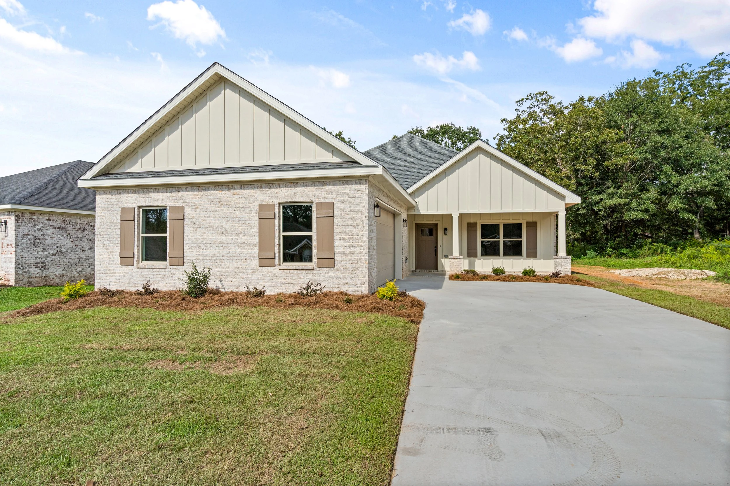 Newly built single-story house with white brick exterior, beige trim, and a two-car garage. Front yard has grass and small shrubs, driveway made of concrete, and surrounded by trees and clear sky.