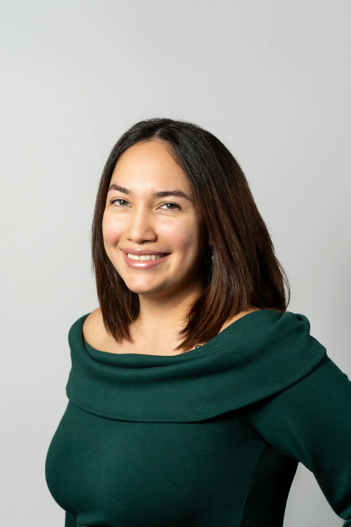 A woman with shoulder-length dark brown hair, smiling, wearing a dark green top with a wide collar, standing against a plain gray background.