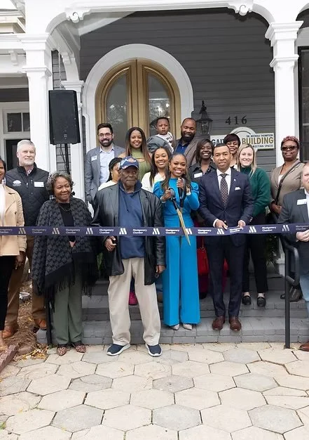 Group of diverse people, including men and women of different ages, gathered on the steps outside a building for a ribbon-cutting ceremony. The woman in the blue suit is holding scissors, preparing to cut the ribbon.