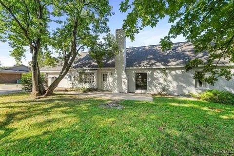 A white house with a dark roof, large trees, and a lush green lawn in the front yard on a sunny day.
