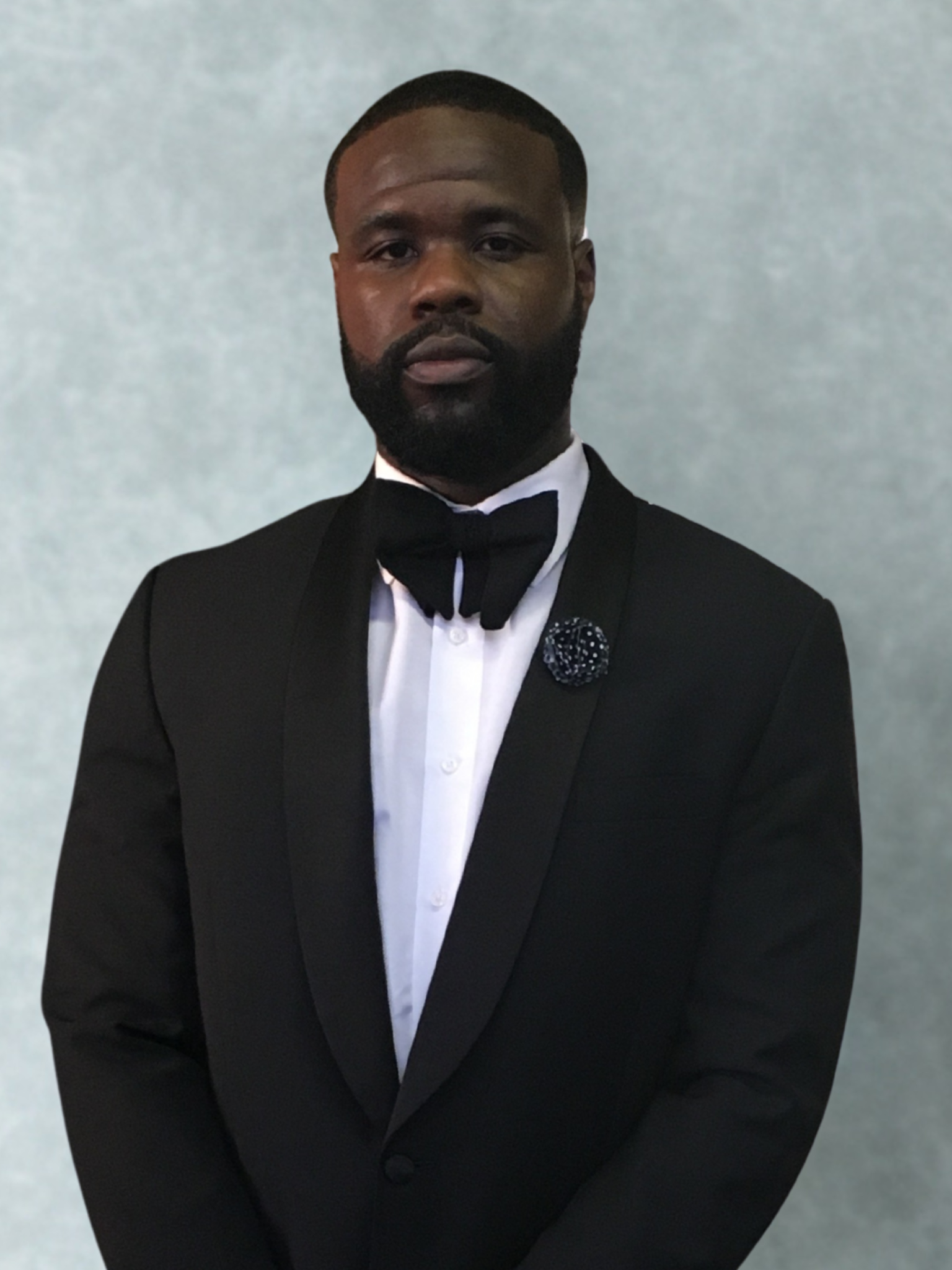 A man in a black tuxedo with a white shirt and black bow tie standing against a light gray background.