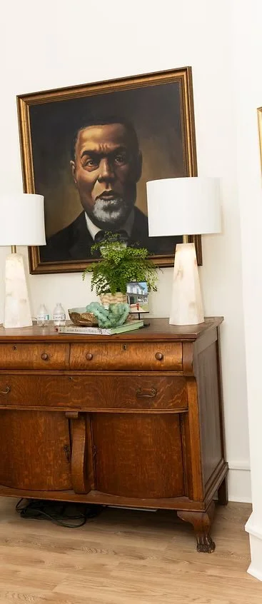 A wooden sideboard with decor and books, topped with two white lamps, in front of a white wall with a large framed black-and-white portrait of a man hanging above.