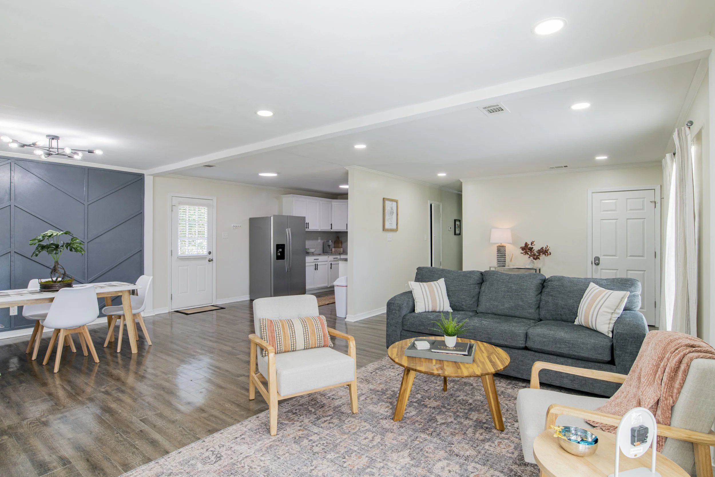 Living room with wooden floors, gray sofa, armchair, round coffee table, dining table with chairs, kitchen area with white cabinets and stainless steel refrigerator, and decorative plants and lamps.