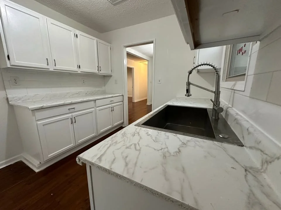 View of a kitchen with white cabinets, marble countertops, and a black sink. There is a doorway leading to another room with hardwood flooring.
