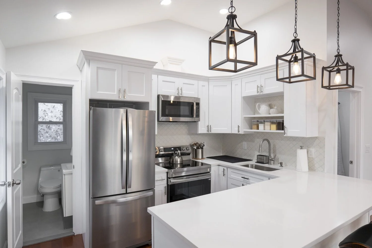 Modern kitchen with white cabinets, stainless steel refrigerator, microwave, and stove. Black pendant lights hang above a white countertop island. Small bathroom with a toilet is visible to the left.