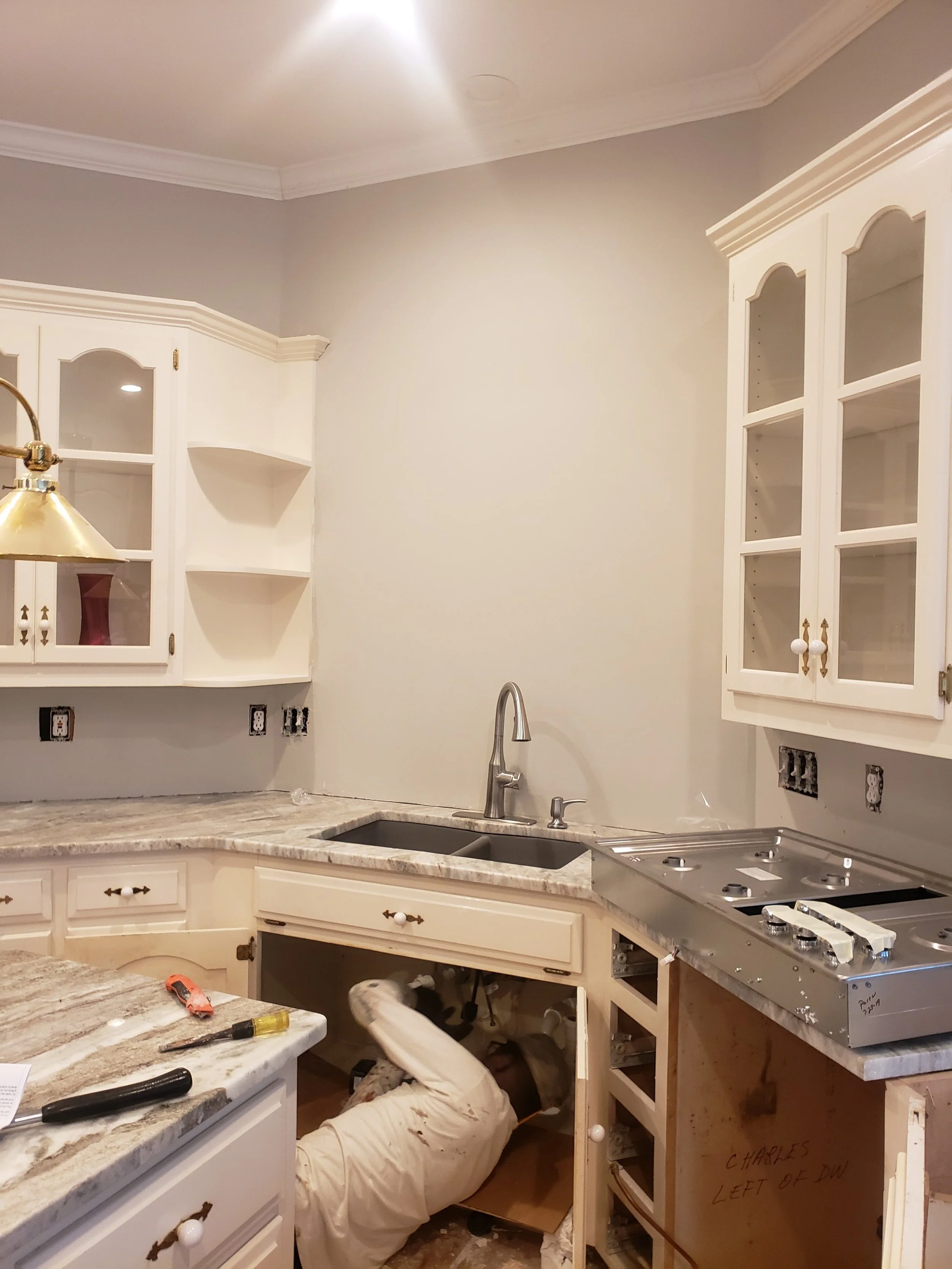 A person working underneath kitchen cabinets during a renovation or repair, with tools and uninstalled cabinet doors visible.