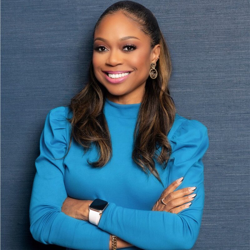 A woman with long brown hair smiling, wearing a blue long-sleeve top, gold earrings, a smartwatch, and rings, standing with arms crossed against a blue textured background.