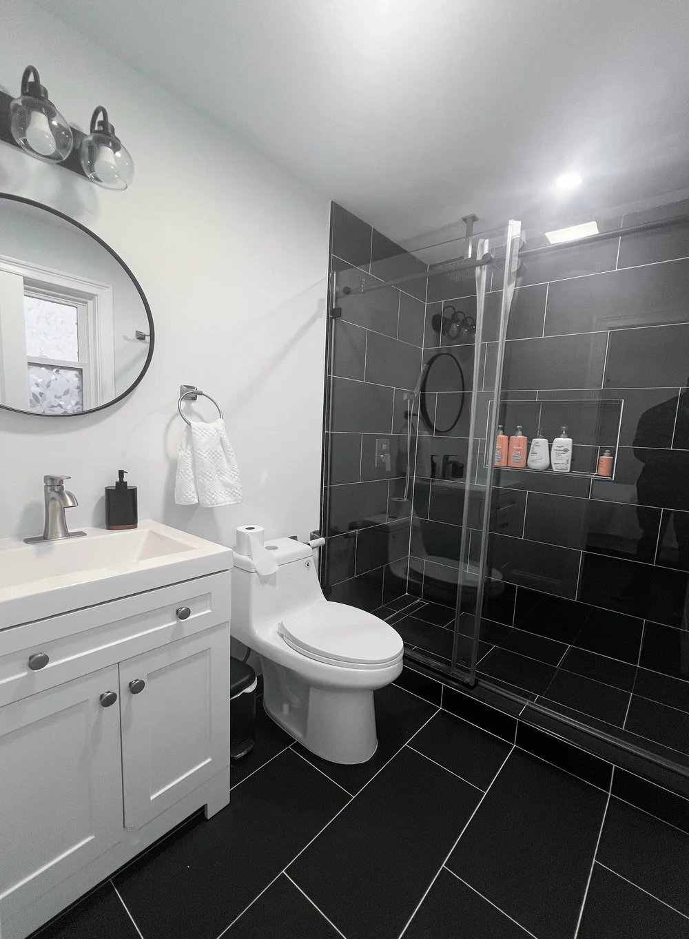 Modern bathroom with white vanity, round mirror, black soap dispenser, white toilet, and a glass-enclosed shower with black tiles and built-in shelf holding skincare products.