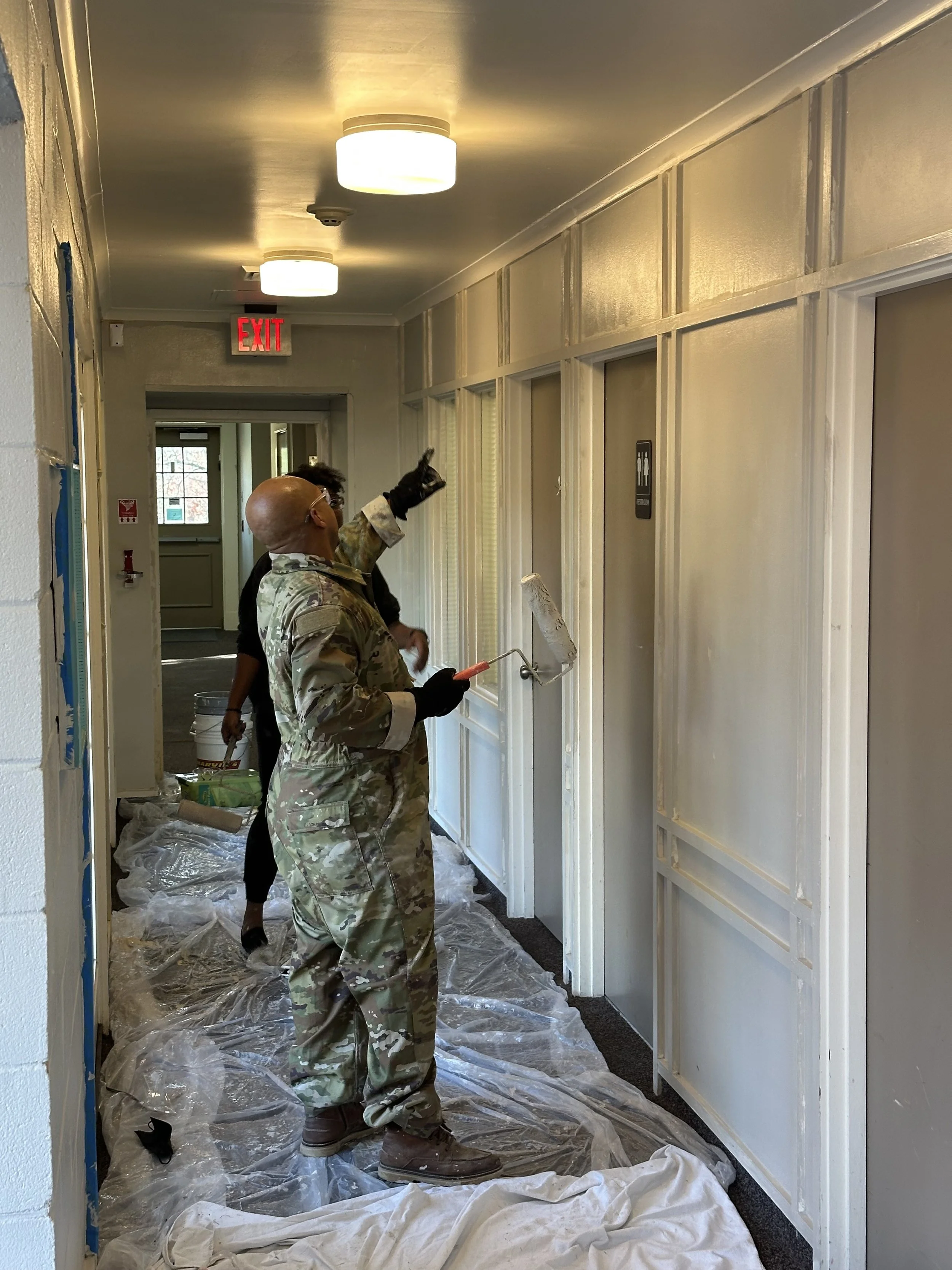 A person in camouflage uniform painting a wall in a hallway with a paint roller, while another person points at the wall. Plastic sheets cover the floor.