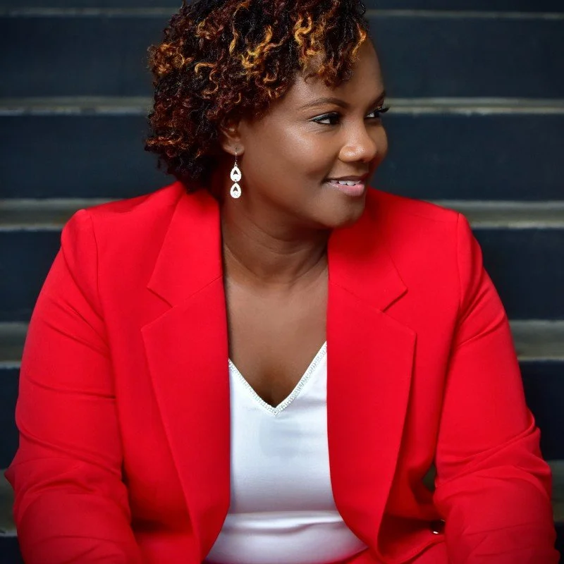 A woman with short curly hair wearing a red blazer and white top, sitting on dark stairs, smiling and looking to her right.