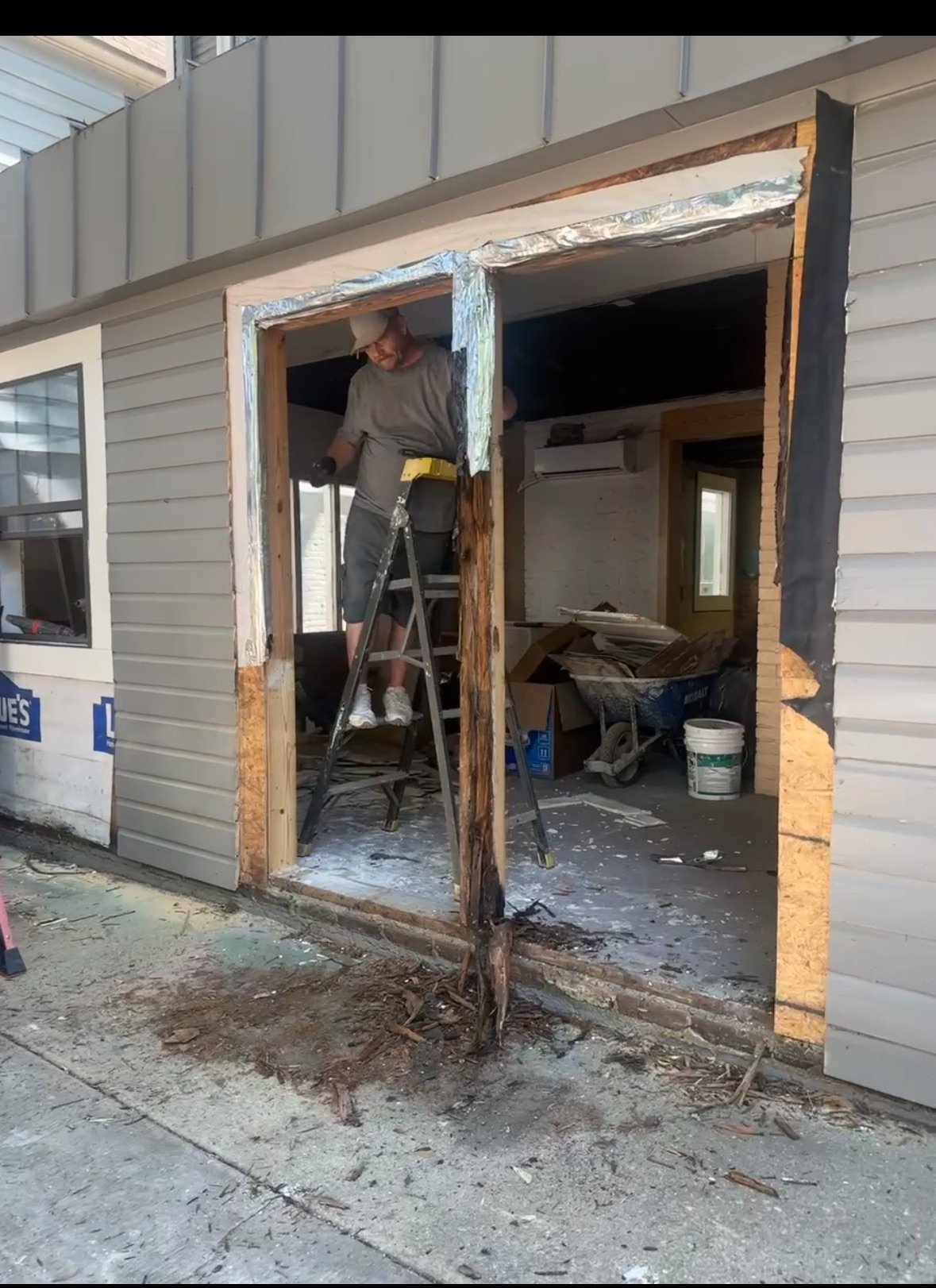 A person working on removing a wall in a house under construction or renovation, standing on a ladder inside, with tools and construction materials around.