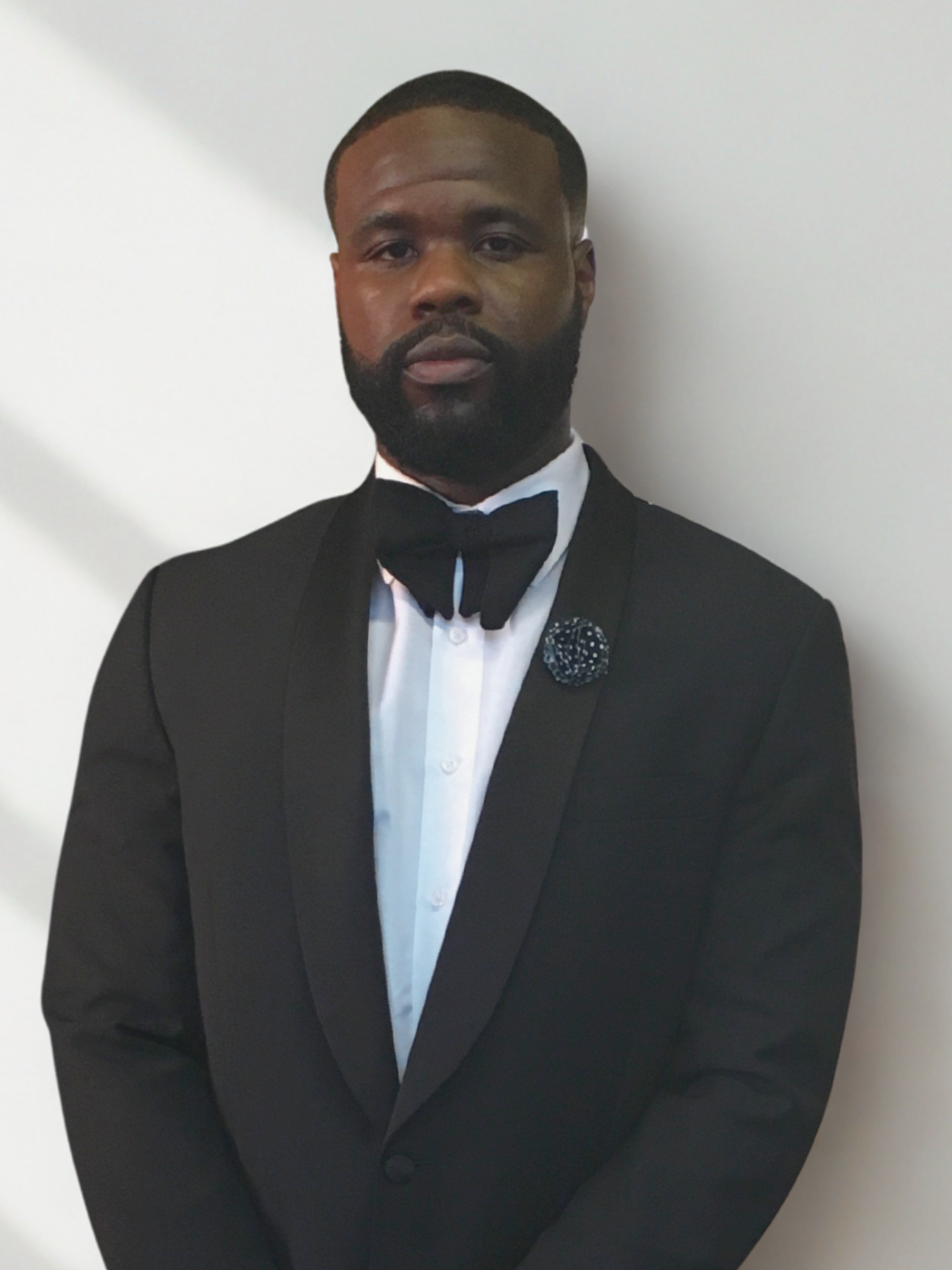 Man dressed in a black tuxedo with a bow tie, standing against a white wall.