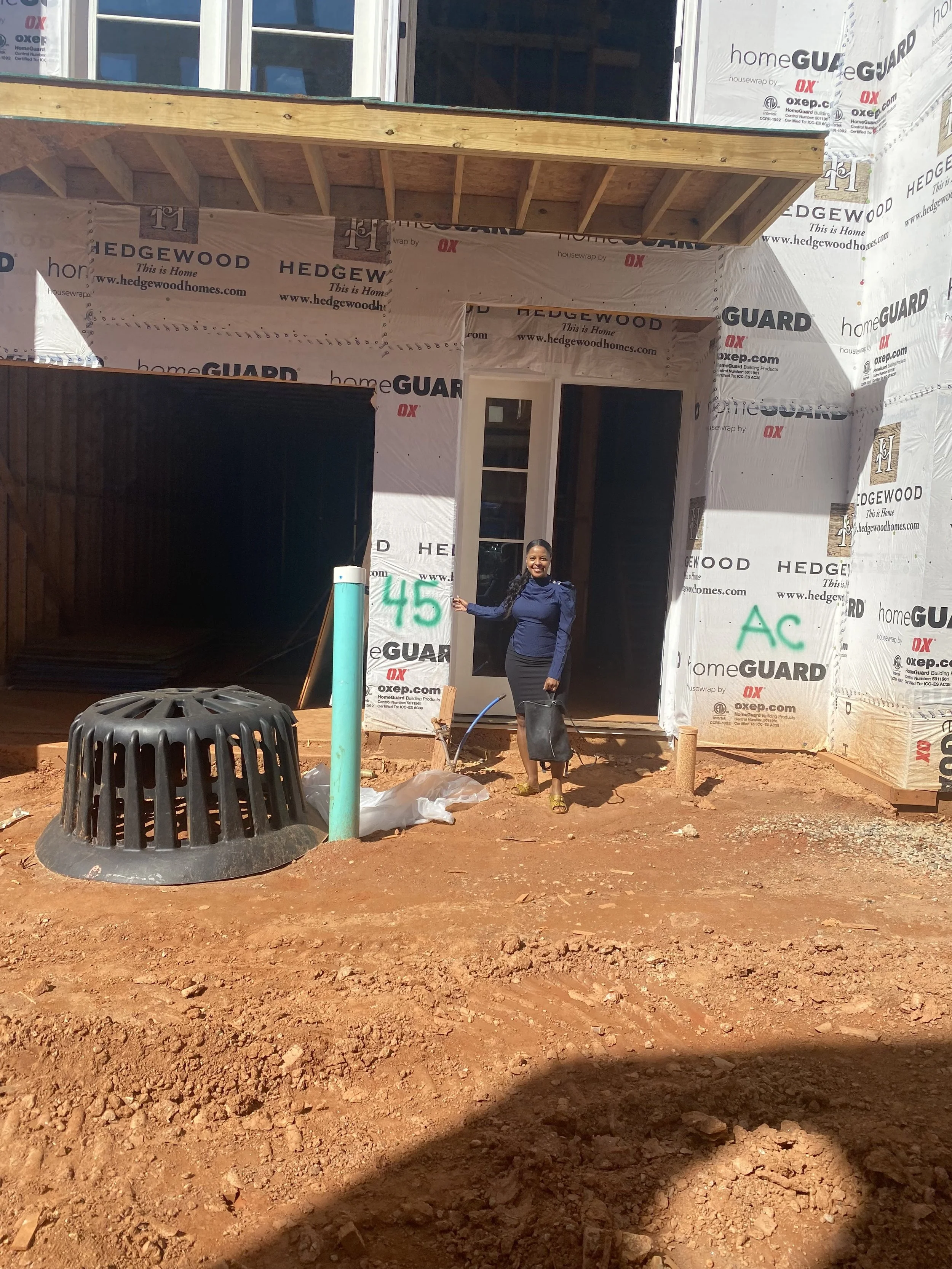 A woman standing in front of a house under construction, giving a thumbs-up. The house has a wooden porch and is wrapped in house wrap with branding. There is a large black drainage grate, a green pipe, and markings '45' and 'AC' on the house wrap. T