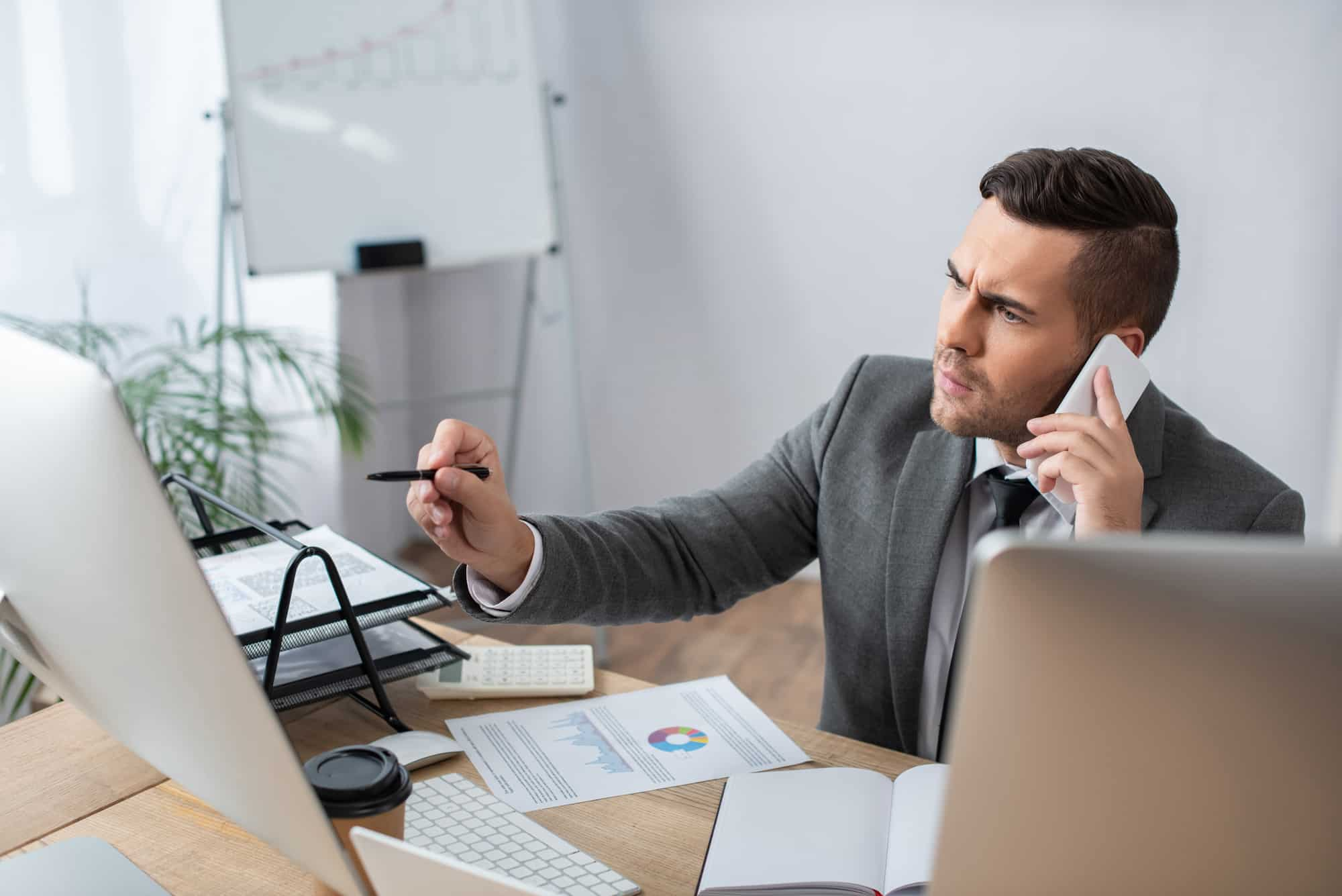 A businessman in a gray suit is sitting at a desk, talking on the phone and holding a pen while looking perplexed at a computer screen.