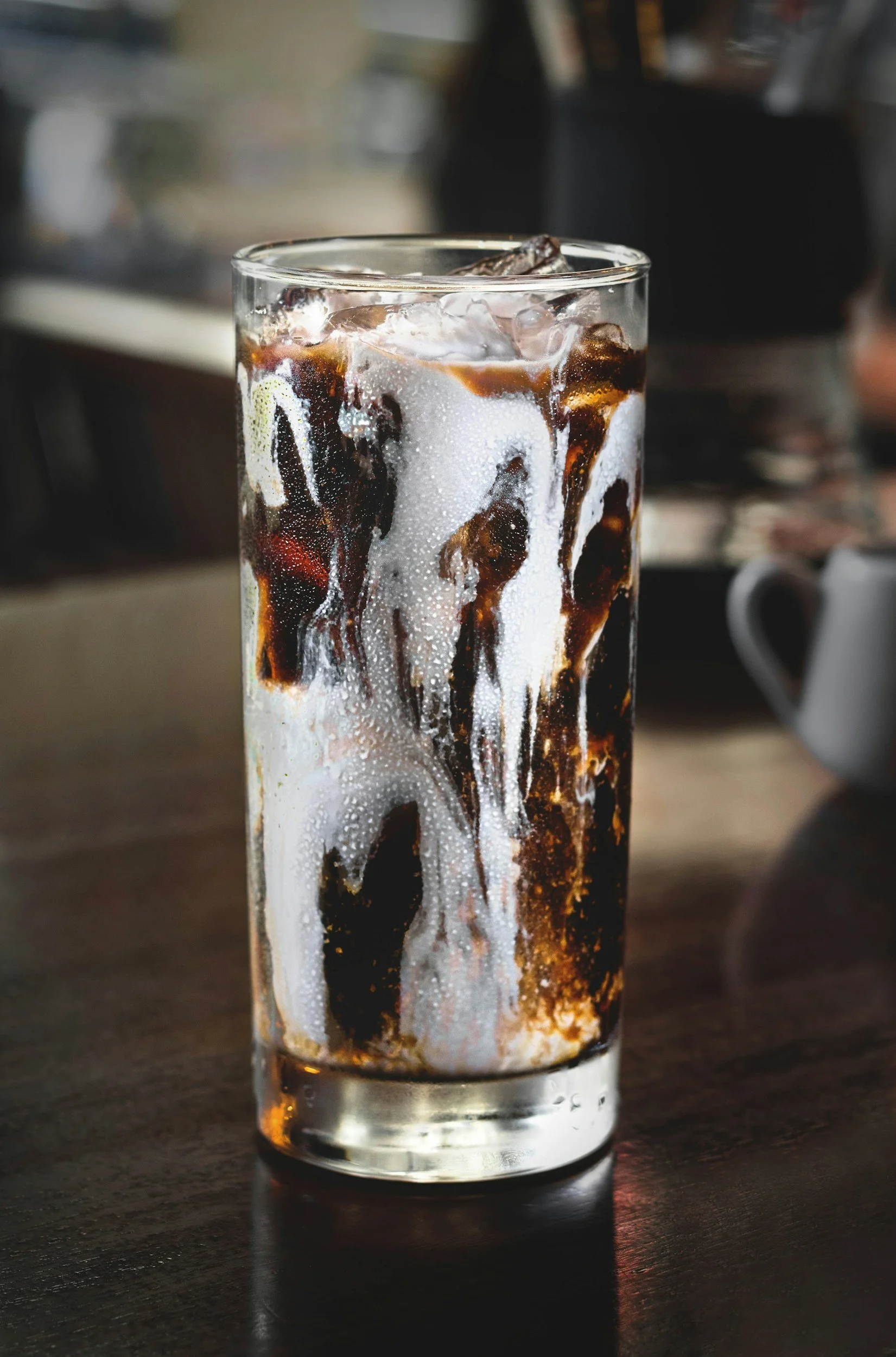 A tall glass of cola with ice and white foam inside, placed on a wooden table in a cafe or restaurant setting.