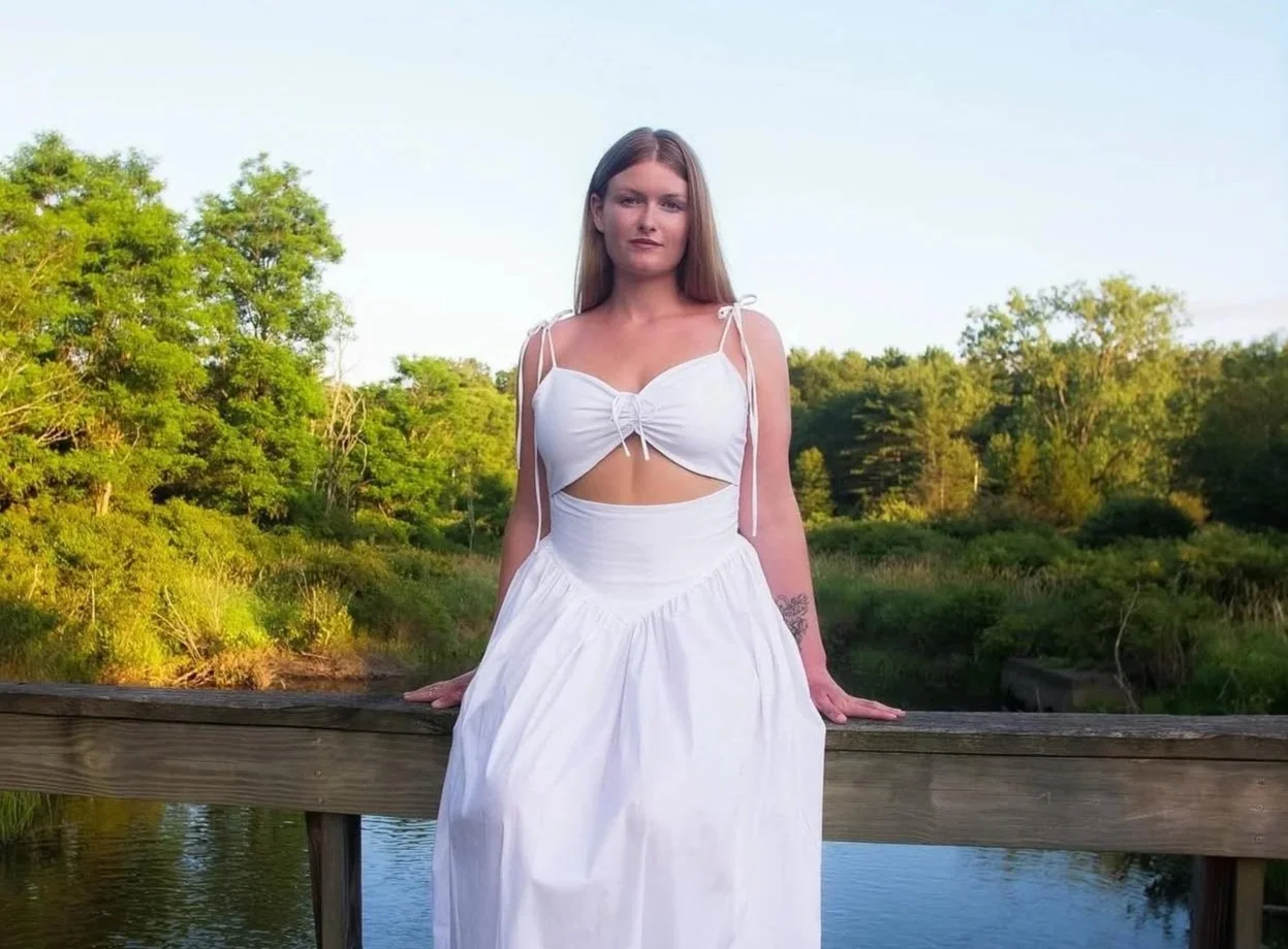 A woman in a white dress with thin straps, standing on a wooden bridge with a river and green trees in the background.
