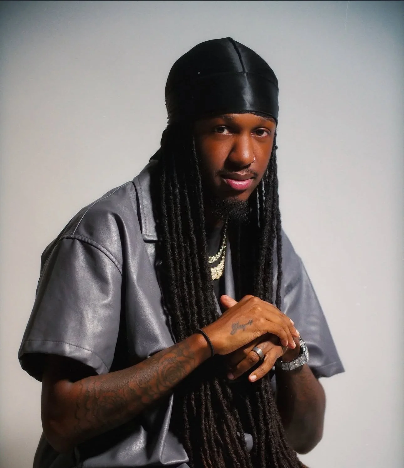 A young man with long dreadlocks, wearing a gray short-sleeve shirt, black bandana, and jewelry, standing against a plain wall.