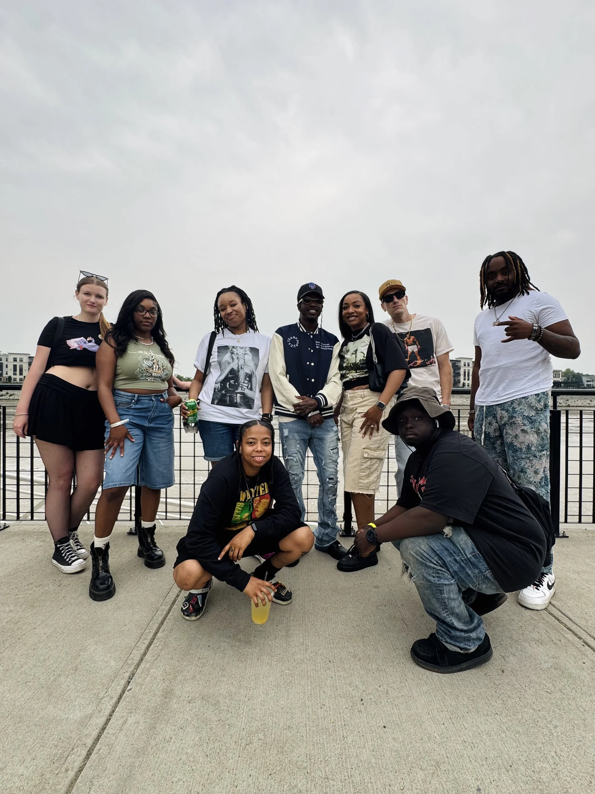 Group of ten young people posing outdoors on a sidewalk area near water with buildings in the background, some standing and two squatting, with casual clothing and accessories.