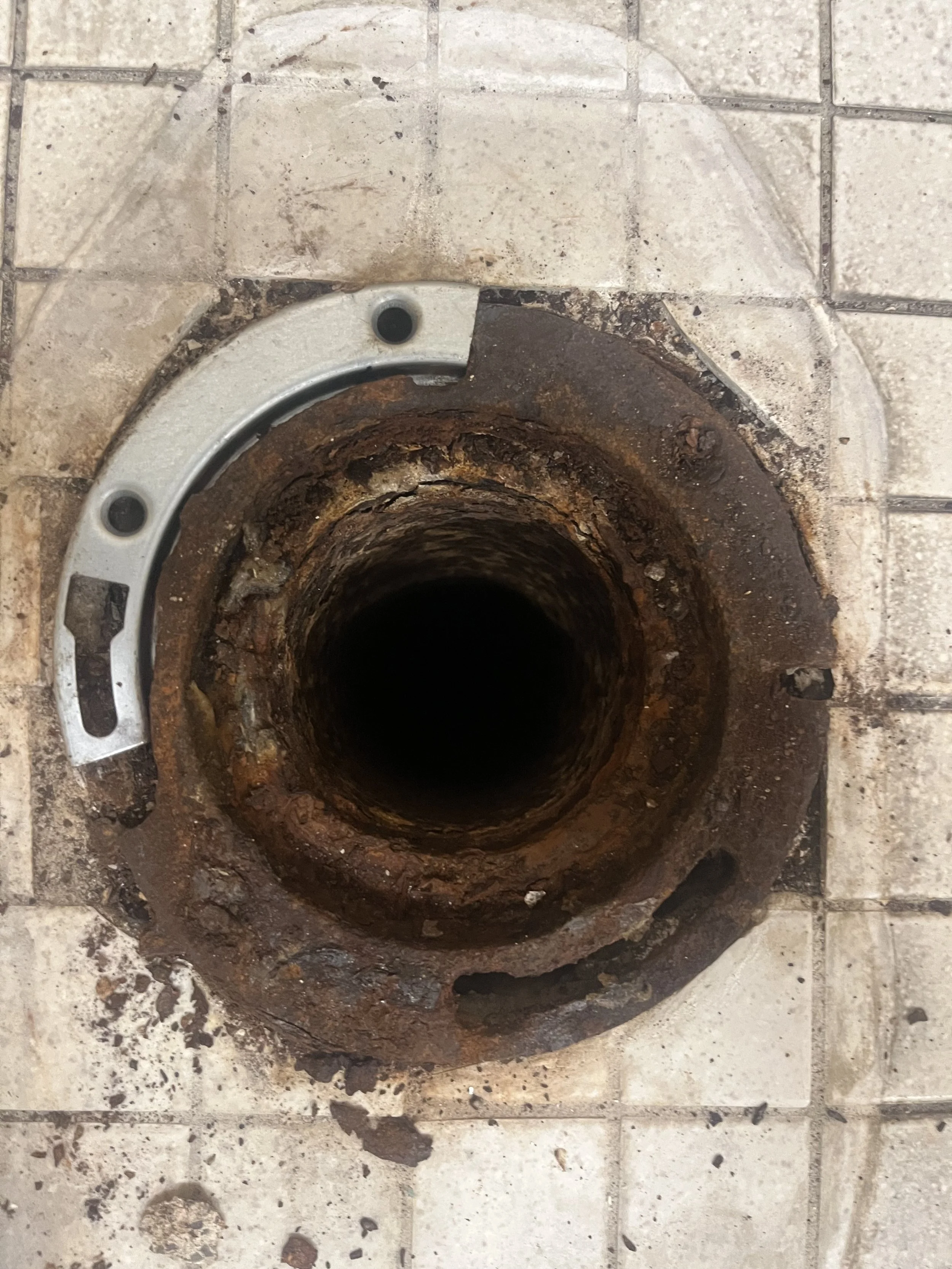 Close-up of a rusty, corroded pipe opening on a tiled floor with a metal flange nearby.