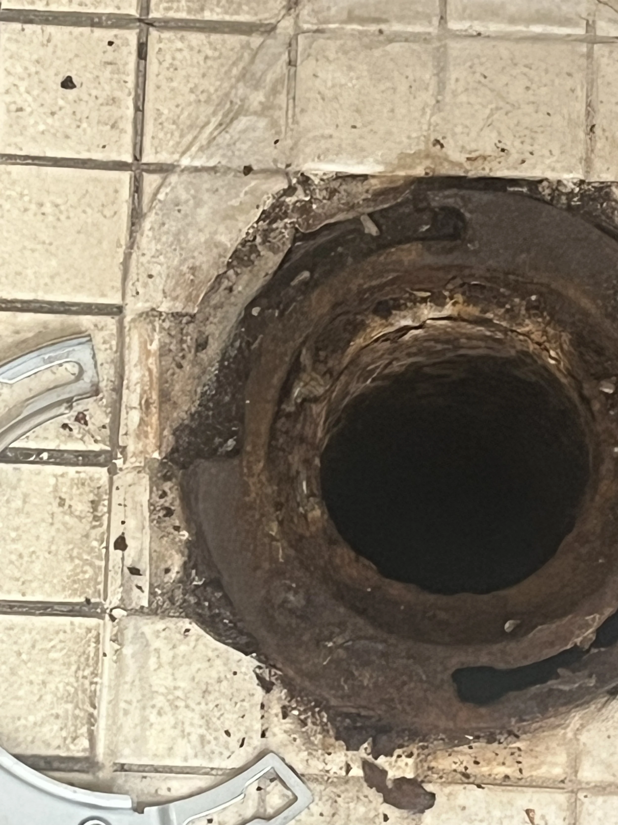 Close-up of a clogged or damaged pipe in a tiled bathroom floor showing rust and buildup around the opening.