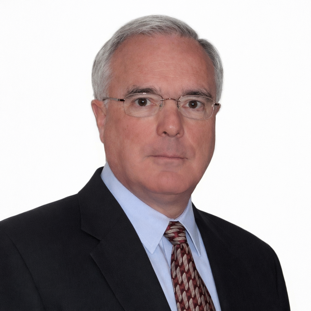 A portrait of a middle-aged man with gray hair and glasses, wearing a dark suit, light blue shirt, and a patterned tie, against a white background.