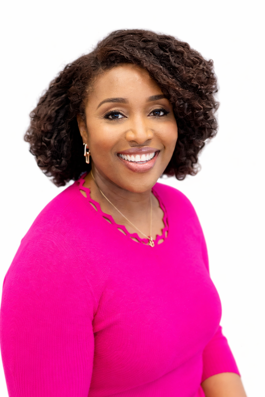 A woman with curly hair, wearing a bright pink top and gold jewelry, smiling against a white background.