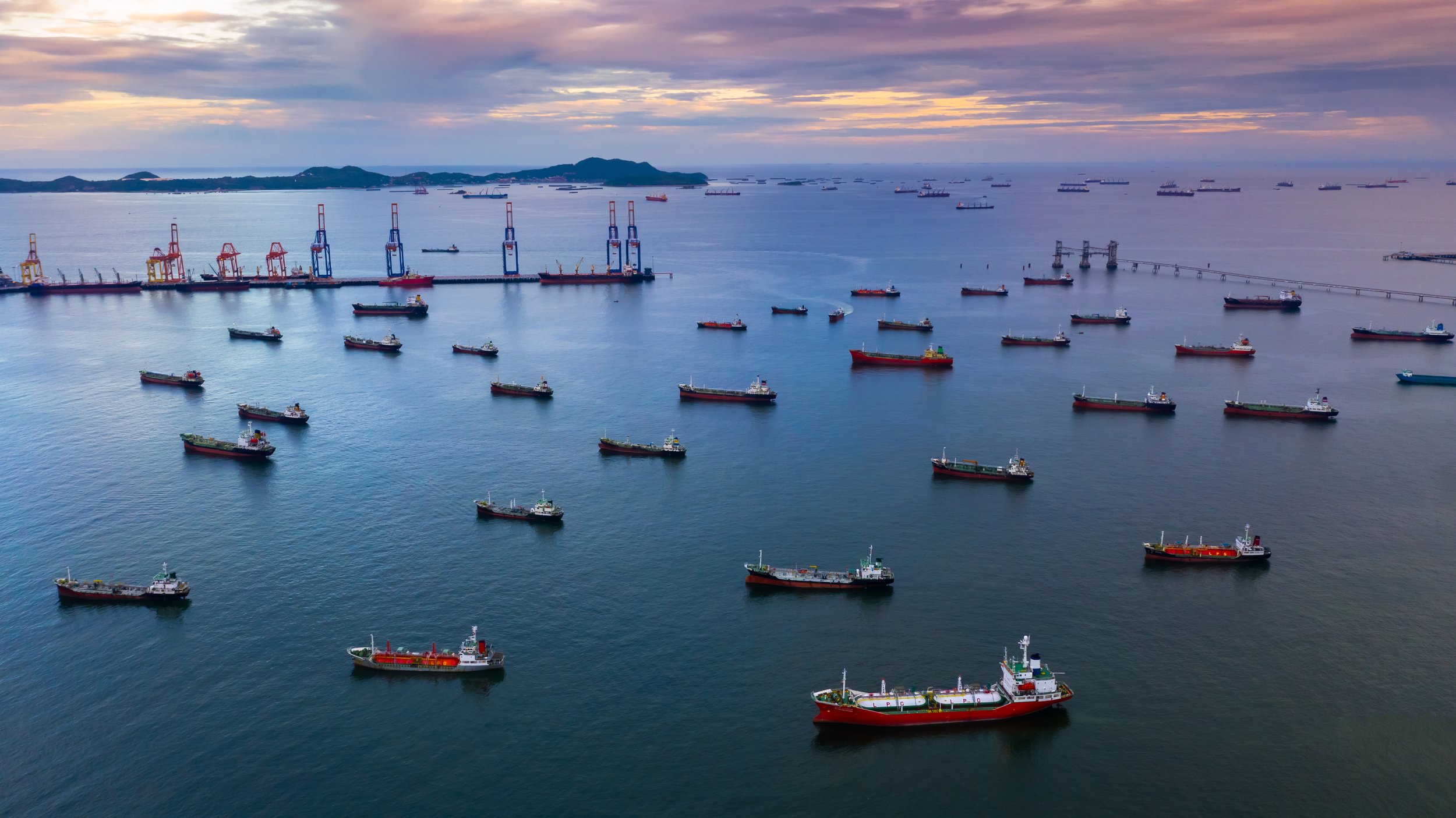 An aerial view of a harbor filled with numerous cargo ships and boats at sea, with a port and cranes in the background. The sky is cloudy with the sun setting or rising.