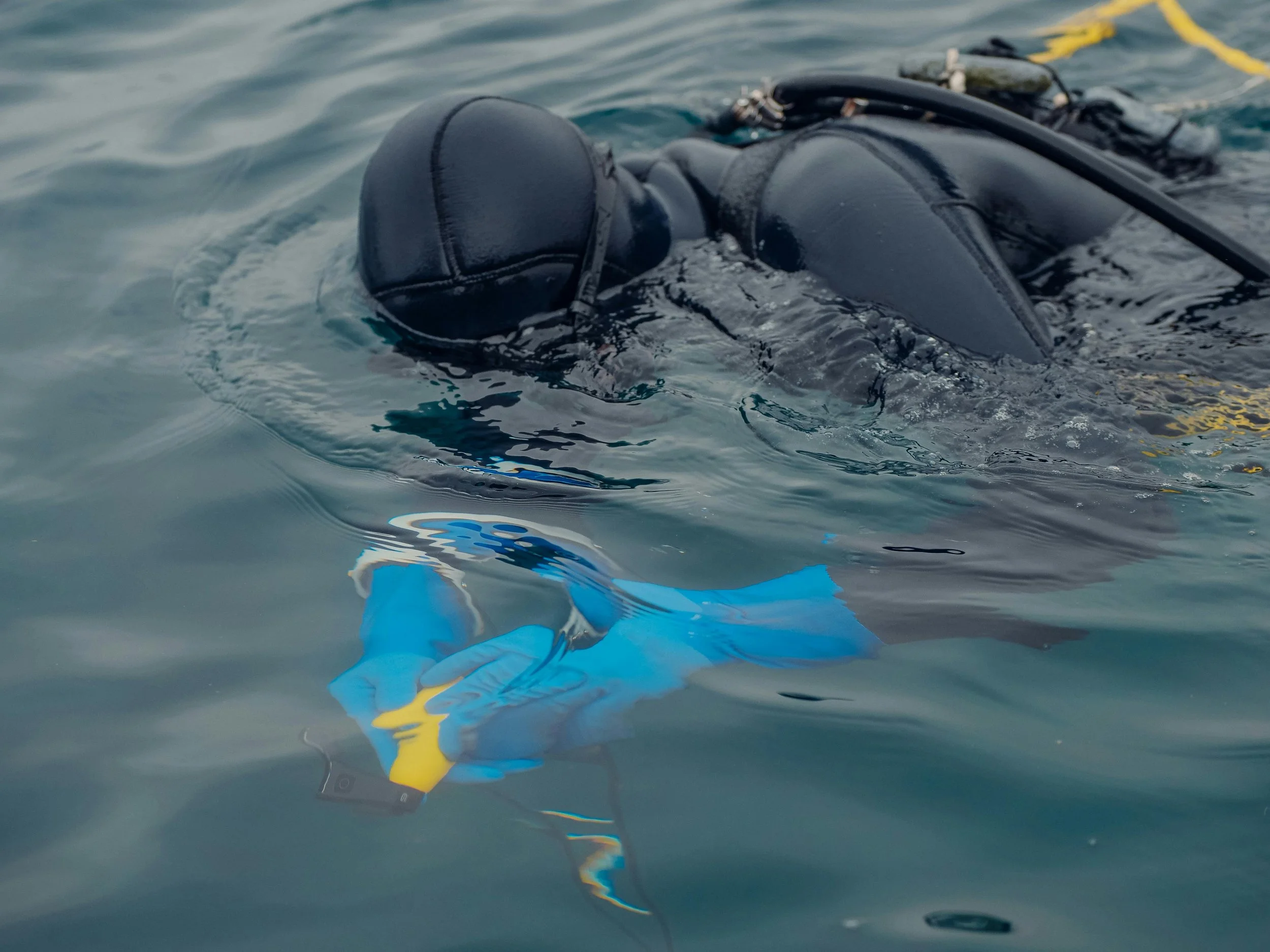 A person in a black wetsuit and diving mask floating face down in the water, holding a waterproof camera in a blue and yellow waterproof case.