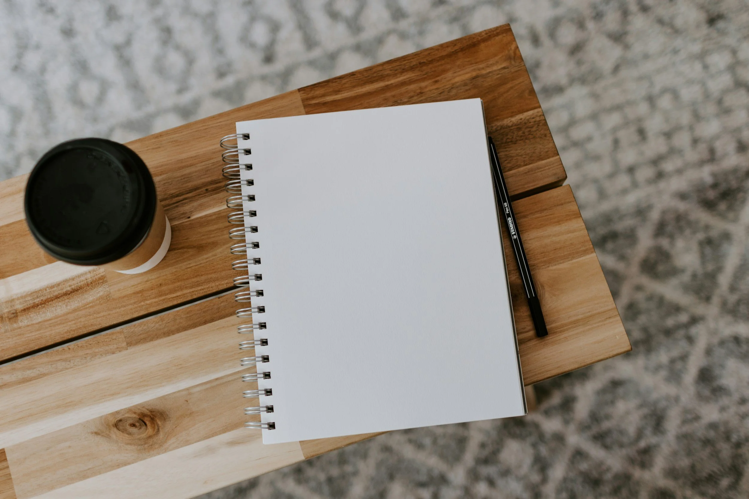 A top-down view of a wooden table with a takeaway coffee cup, a spiral notepad with blank pages, and a black gel pen on a gray textured surface.